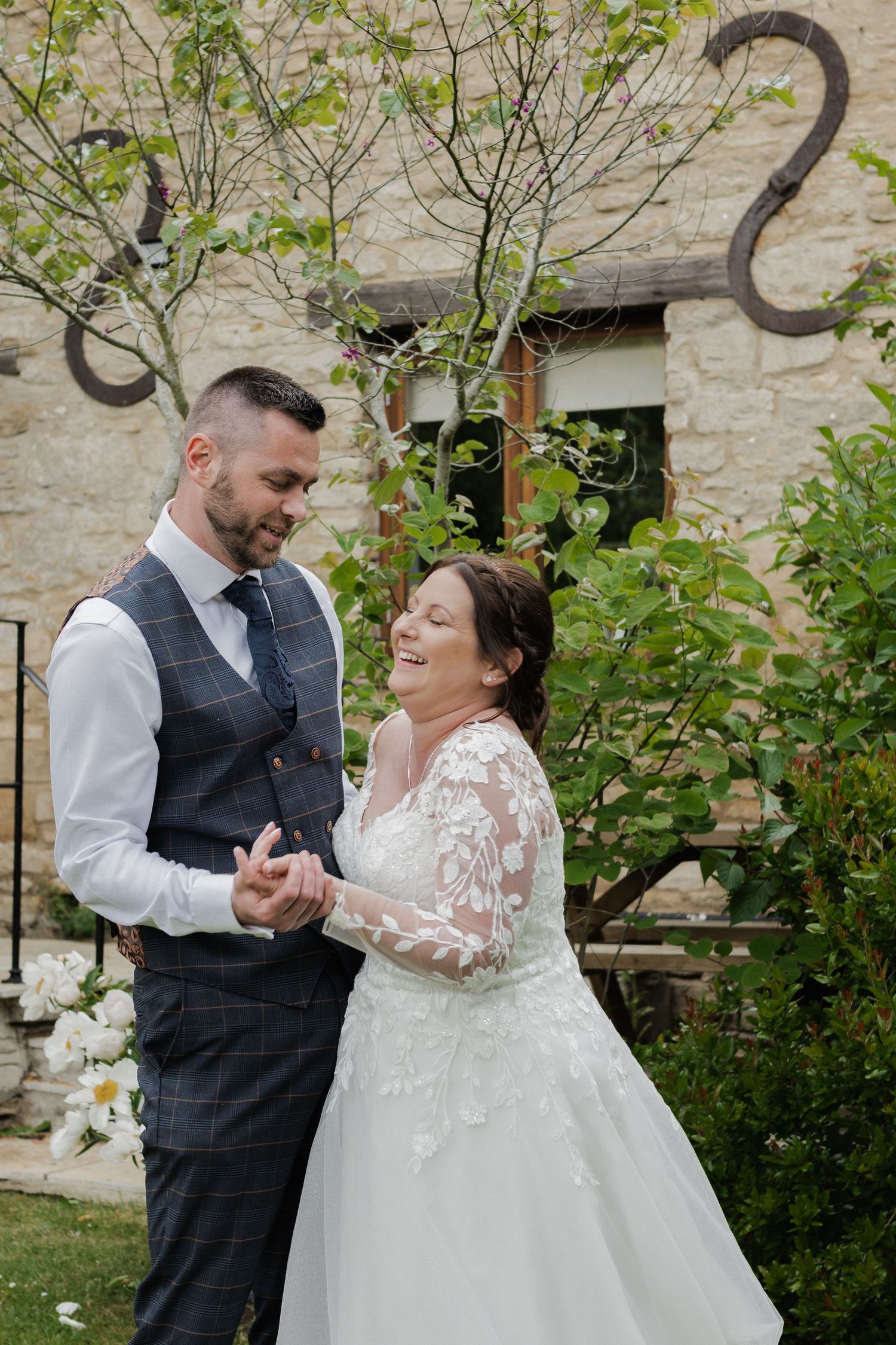 Laughing bride in lace gown holding hands with groom outside rustic stone building.