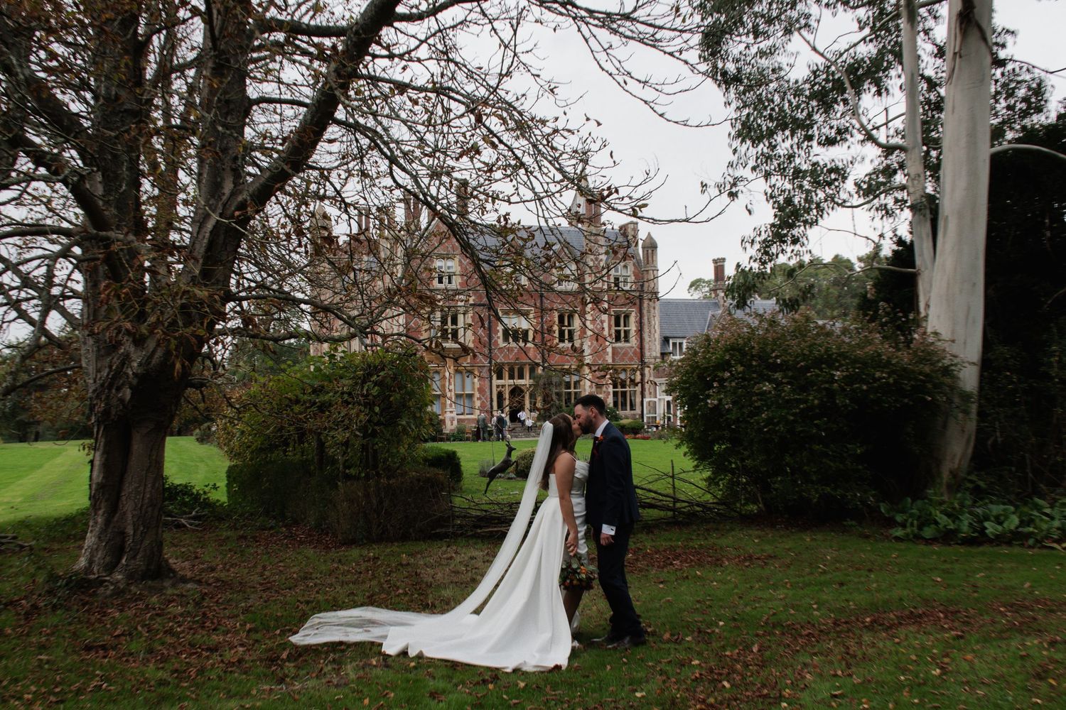 Bride and groom standing together on lush grounds with a stunning historic manor house in the background.