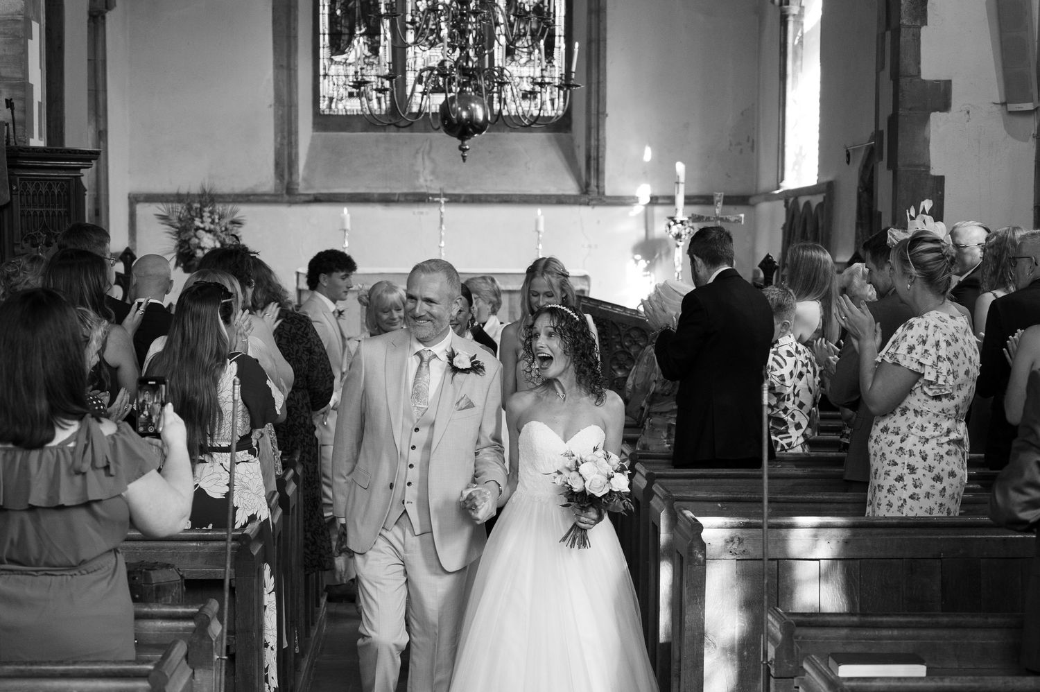 Newlyweds walk back down church aisle together as guests celebrate in black and white photo.