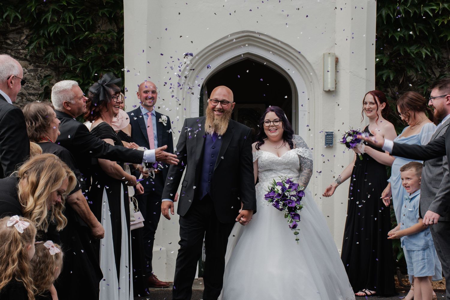 Joyful newlyweds walking through confetti shower outside historic stone church with wedding guests.