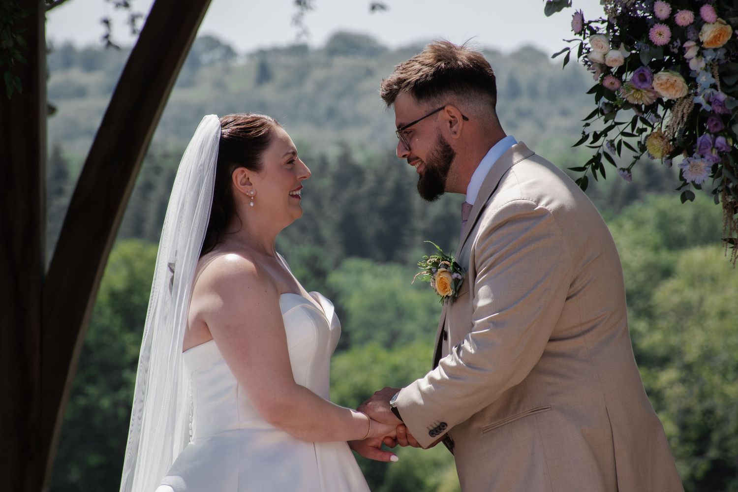Bride and groom holding hands at outdoor wedding ceremony with floral arch and lush green trees in background.
