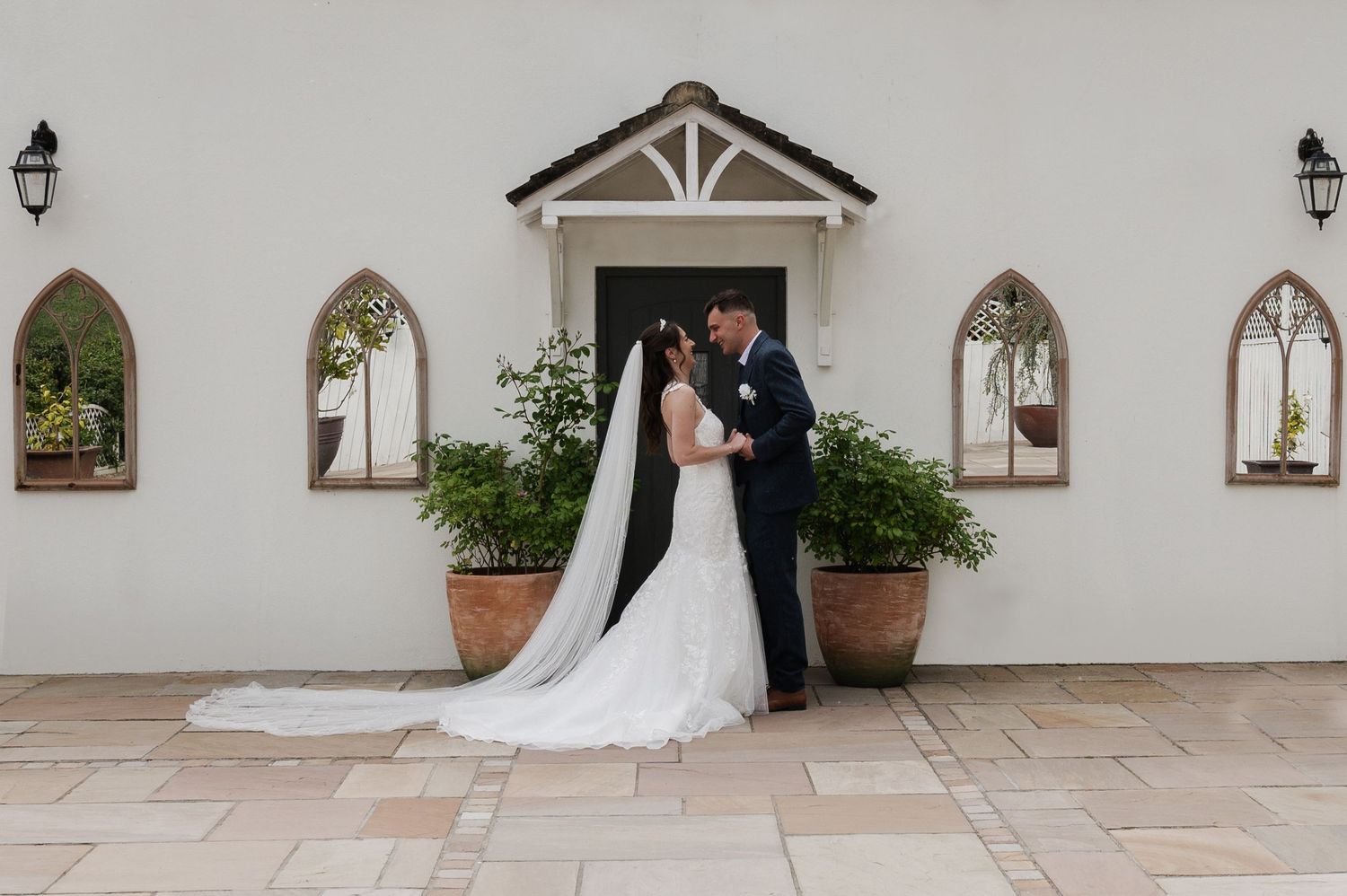 Bride and groom share a romantic moment outside a white chapel with arched windows and lush greenery on their wedding day.