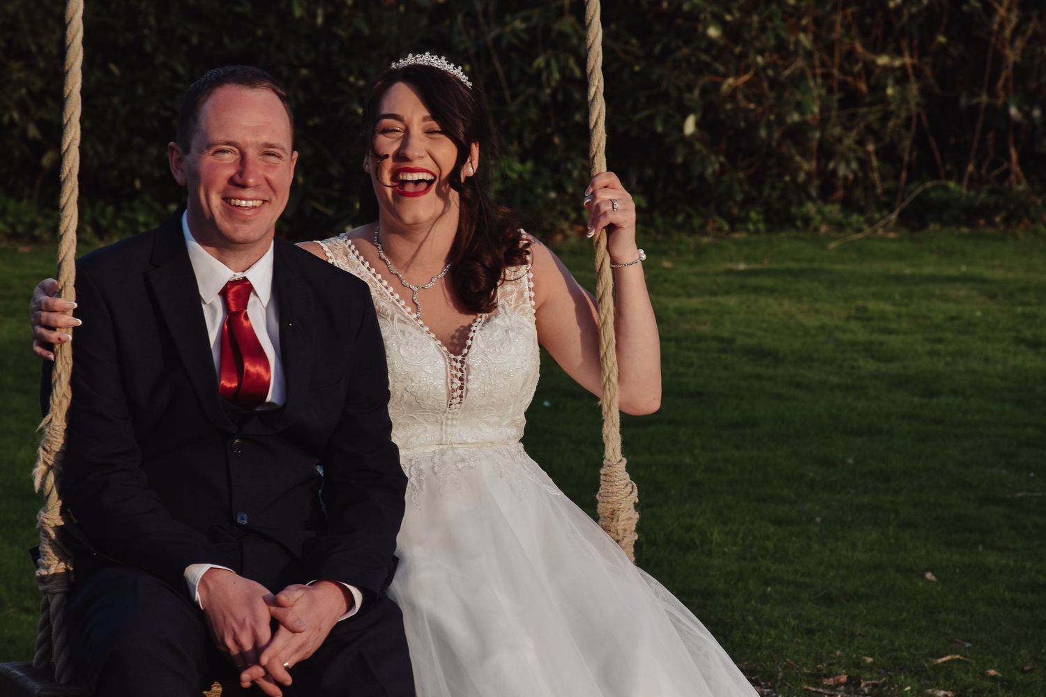 Laughing bride and groom sitting on a rope swing in a garden on their wedding day.