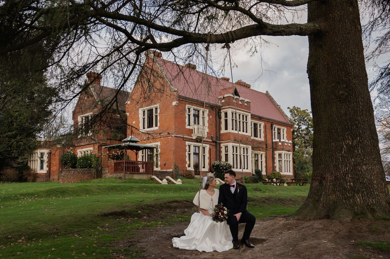 Bride and groom seated together beneath bare tree outside a grand red brick English country house venue.