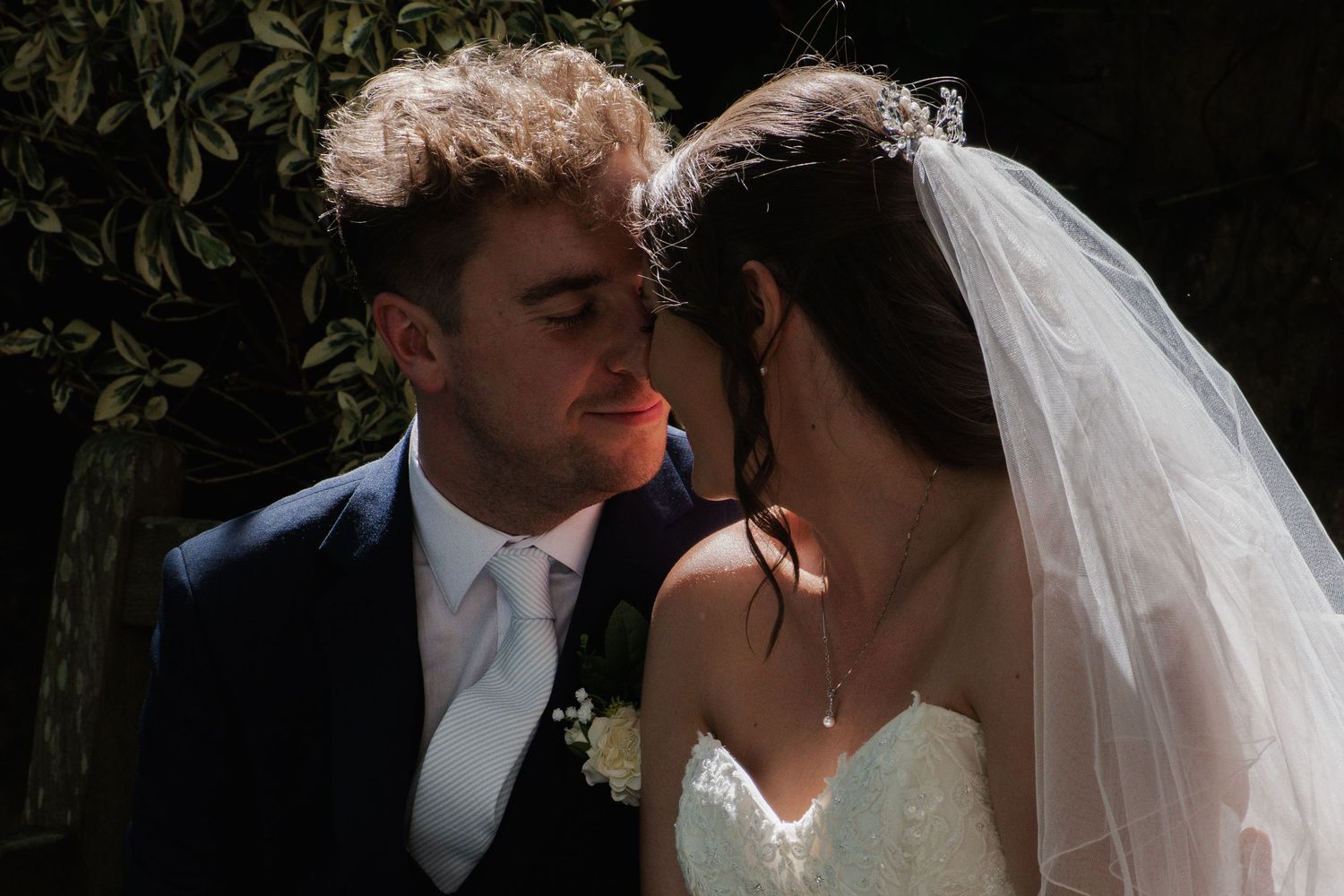 Close-up of newlywed couple sharing an intimate moment, bride wearing veil and groom in navy suit surrounded by greenery.