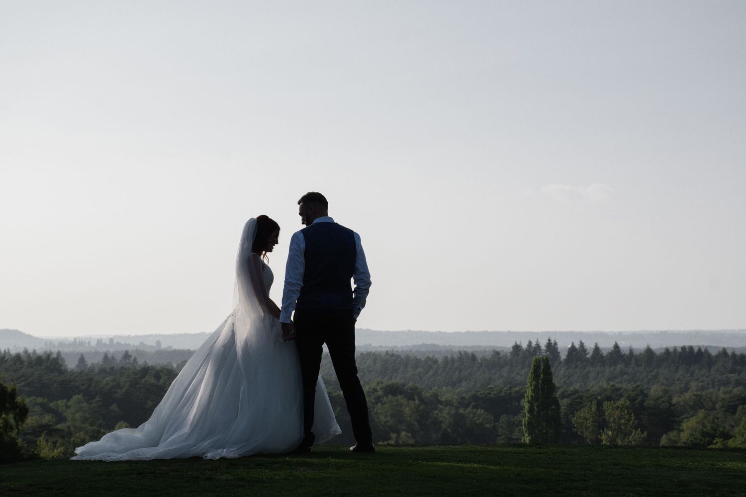Silhouetted bride and groom hold hands overlooking lush green landscape at golden hour.