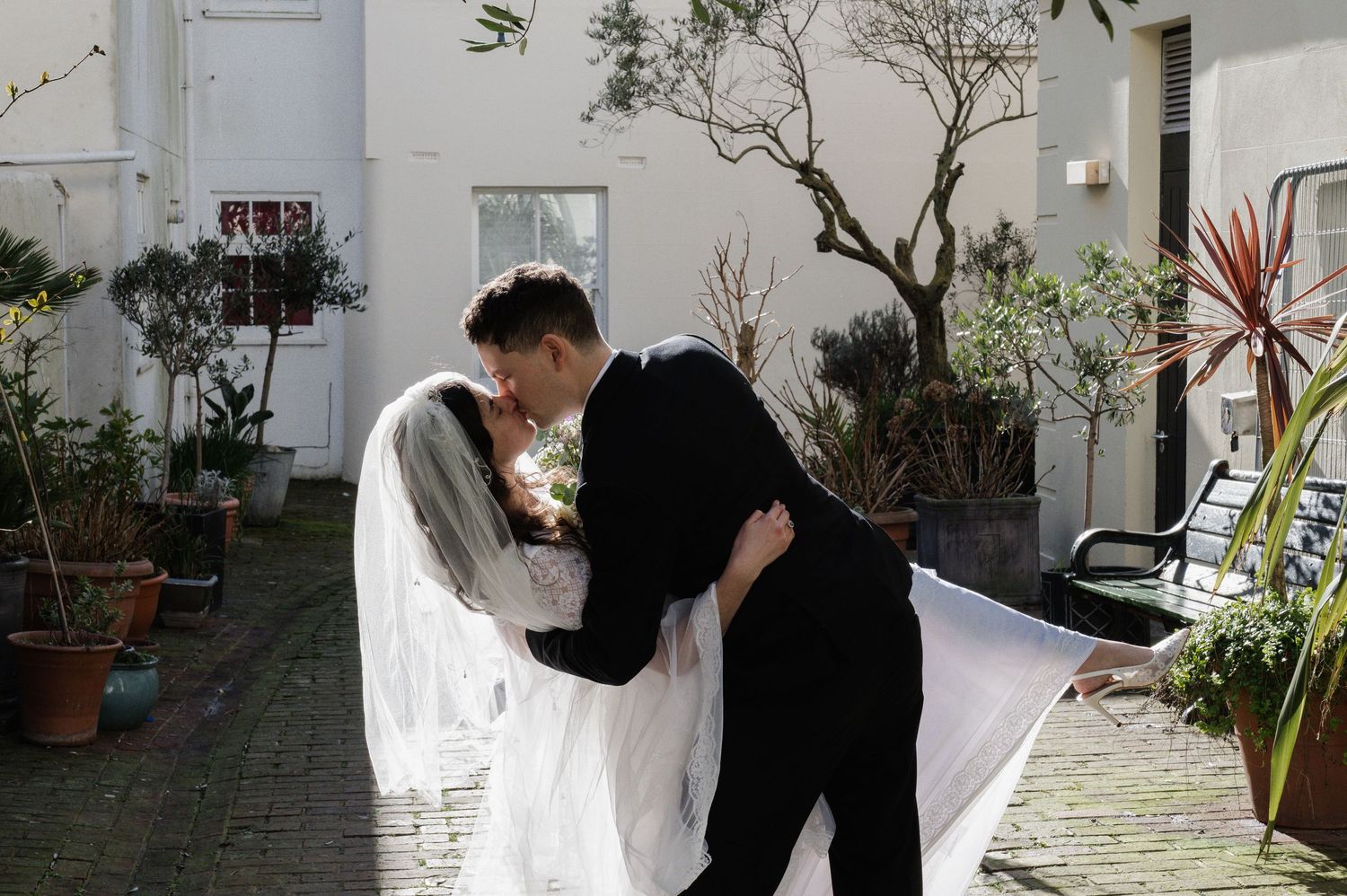 Bride and groom sharing a romantic dip kiss in a charming courtyard with potted plants and white walls.
