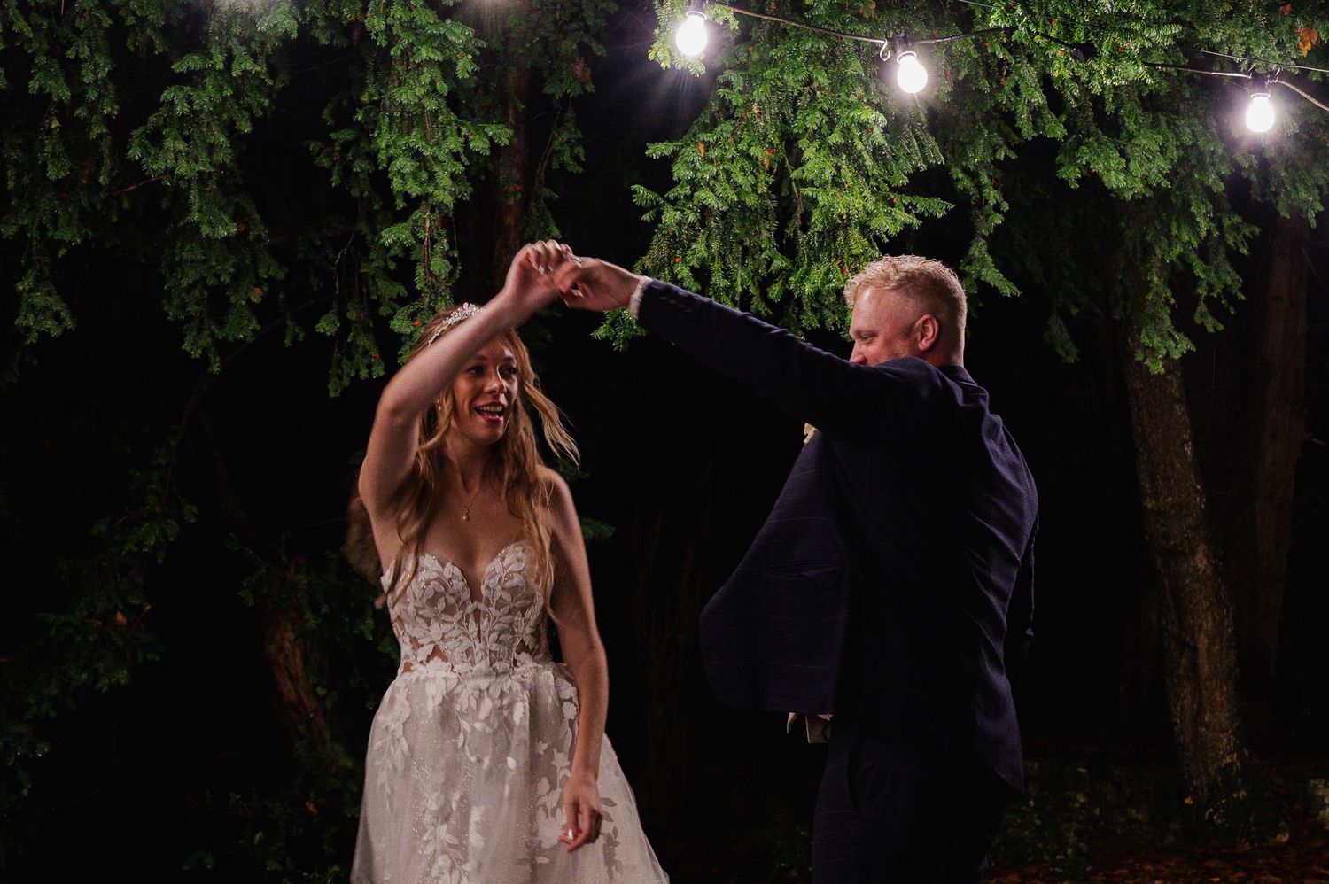 Bride and groom dancing outdoors at night under string lights and lush green foliage at their wedding reception.