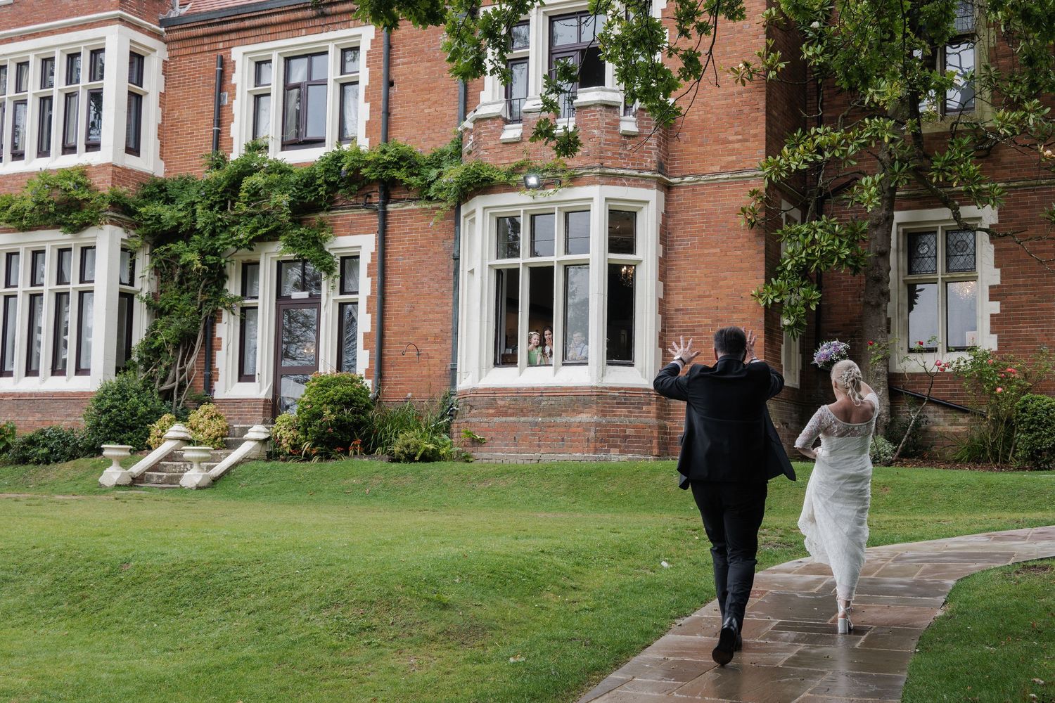 Bride and groom walking hand in hand towards a grand red brick manor house surrounded by lush green gardens.