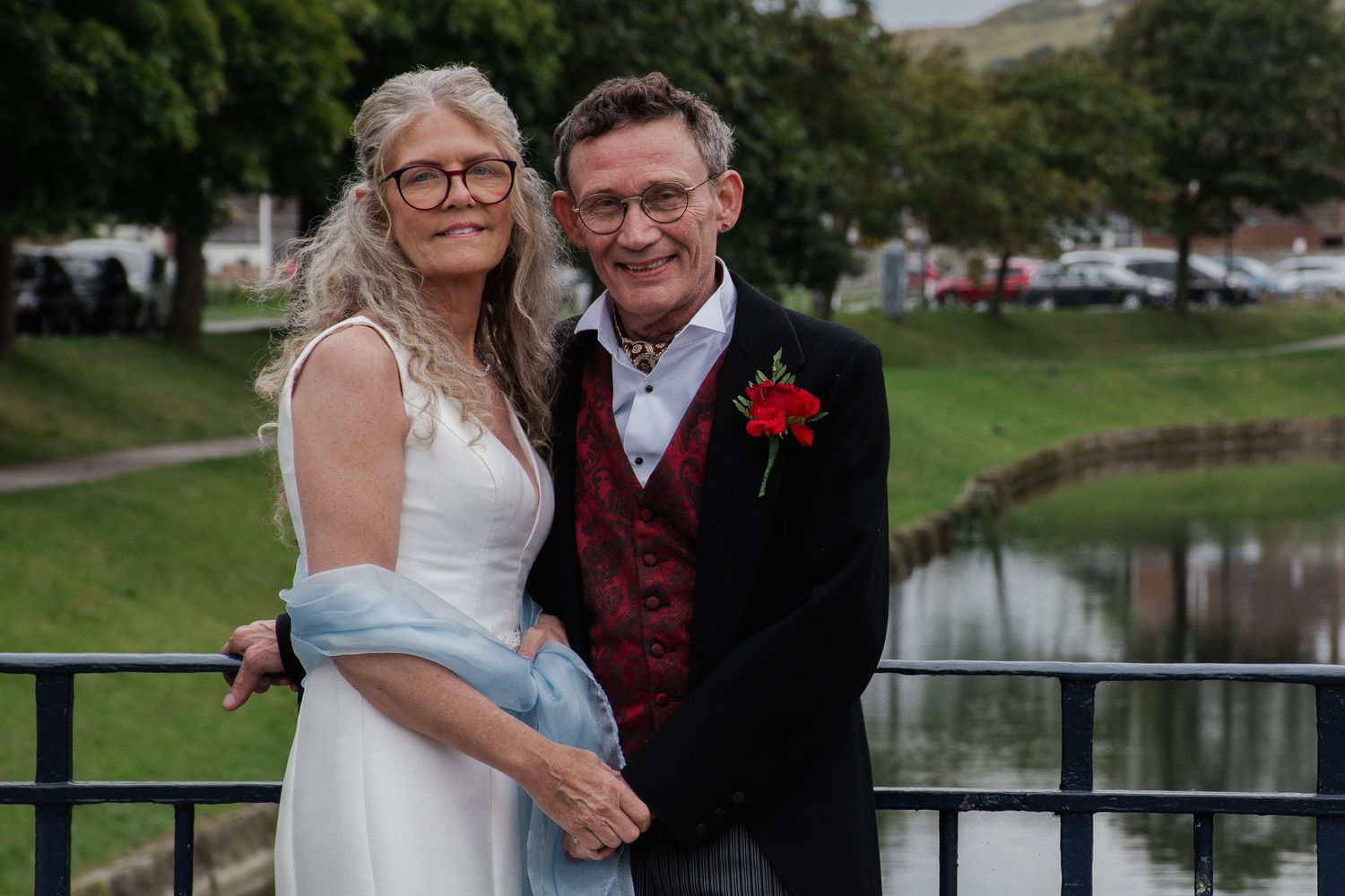 Mature wedding couple smile together on a bridge over a river, bride in white with blue wrap, groom in black with red rose.