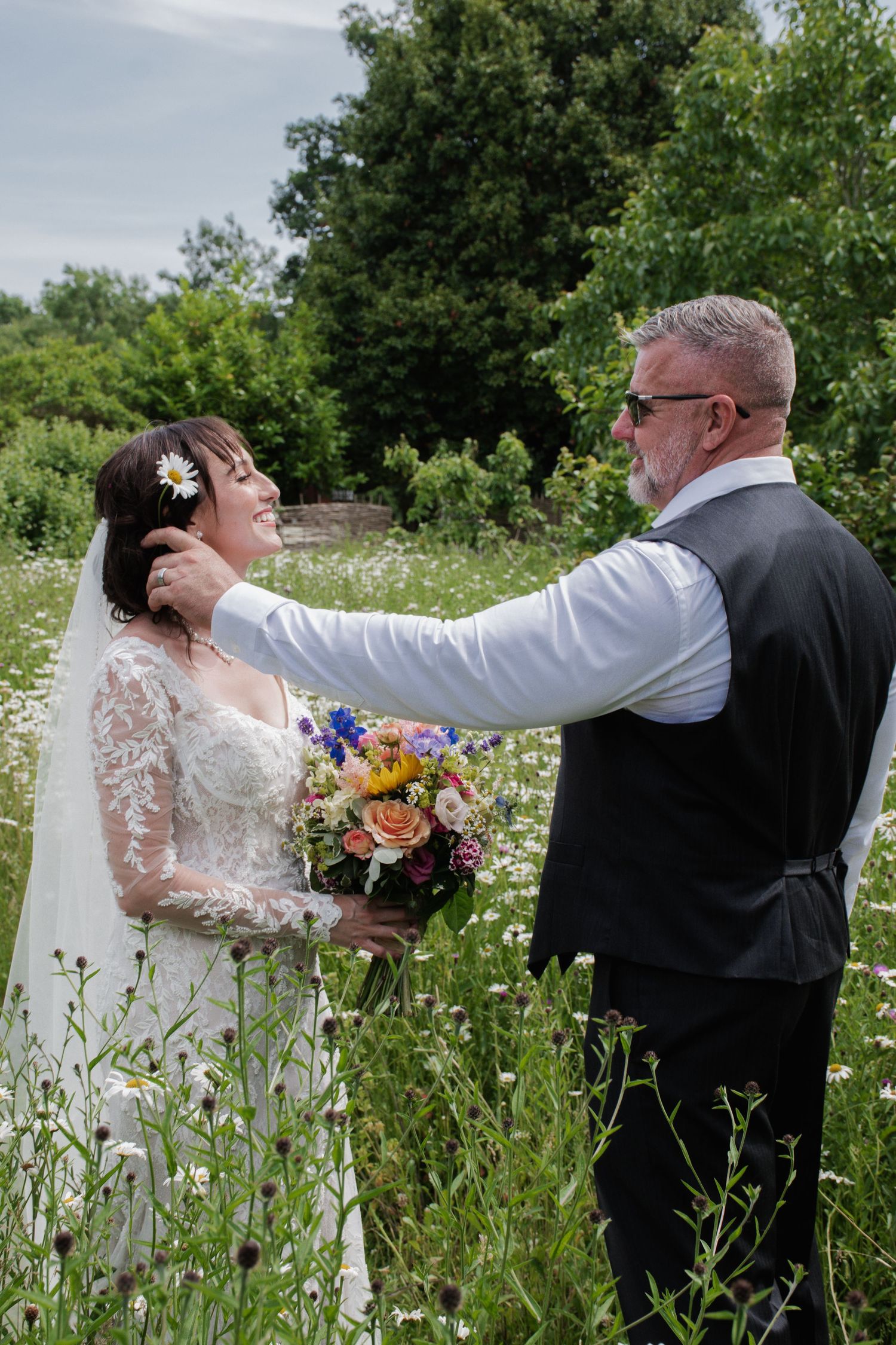 Groom tenderly places flower in brides hair while standing in a wildflower meadow together.