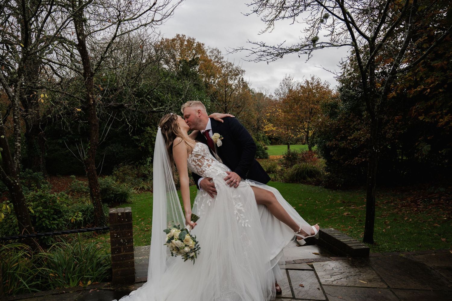 Bride and groom sharing a romantic dip kiss in an autumn garden on their wedding day.