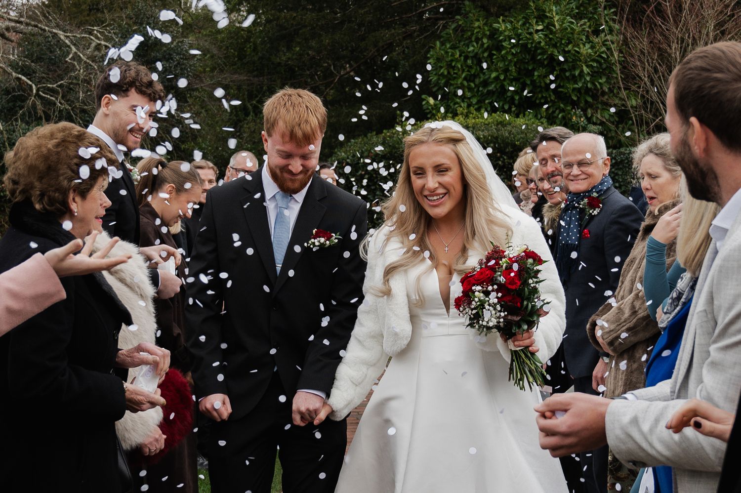 Newlyweds walk through confetti shower, bride holding red roses bouquet, guests celebrating outdoors.