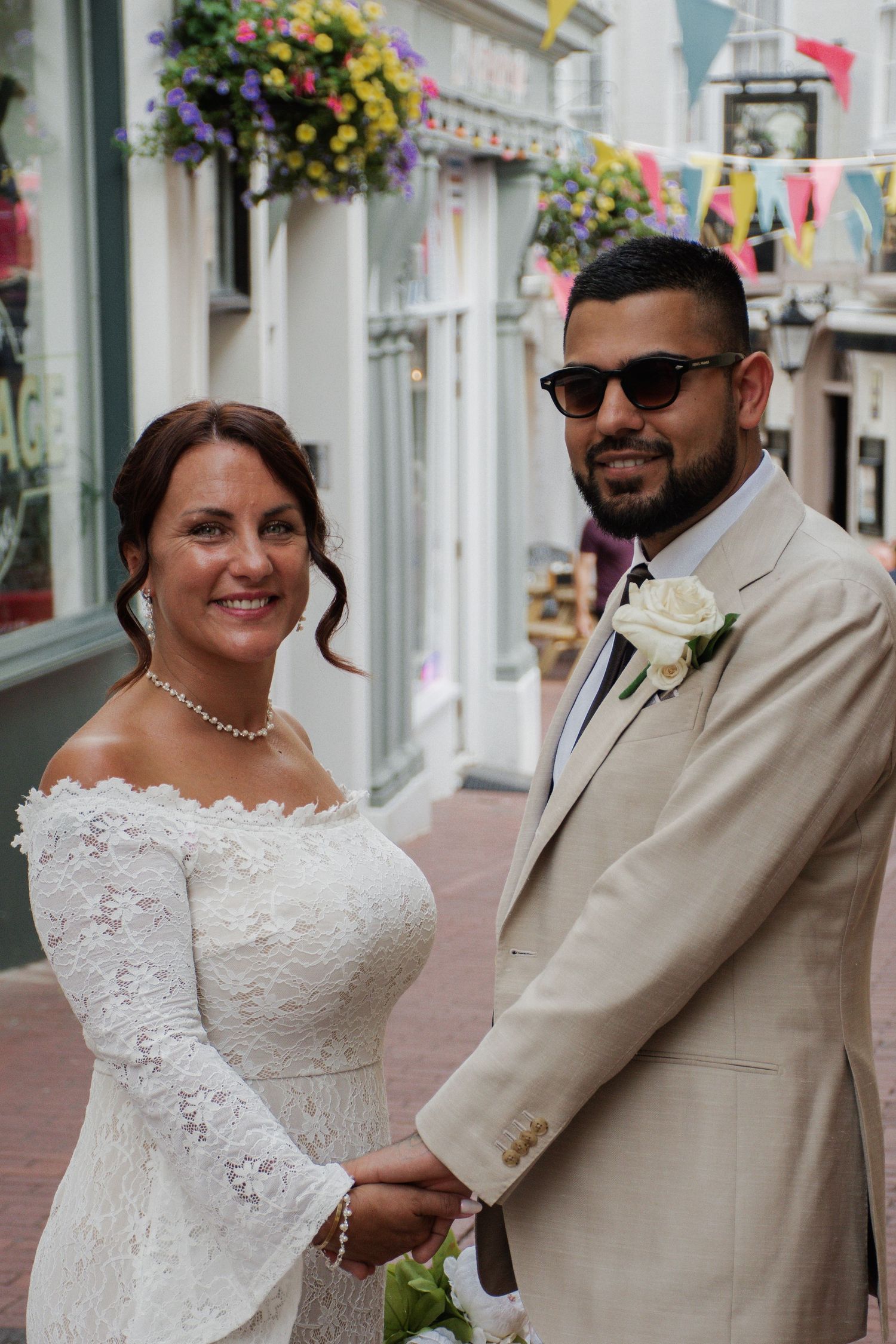 Smiling bride in off-shoulder lace dress and groom in beige suit holding hands on decorated street.