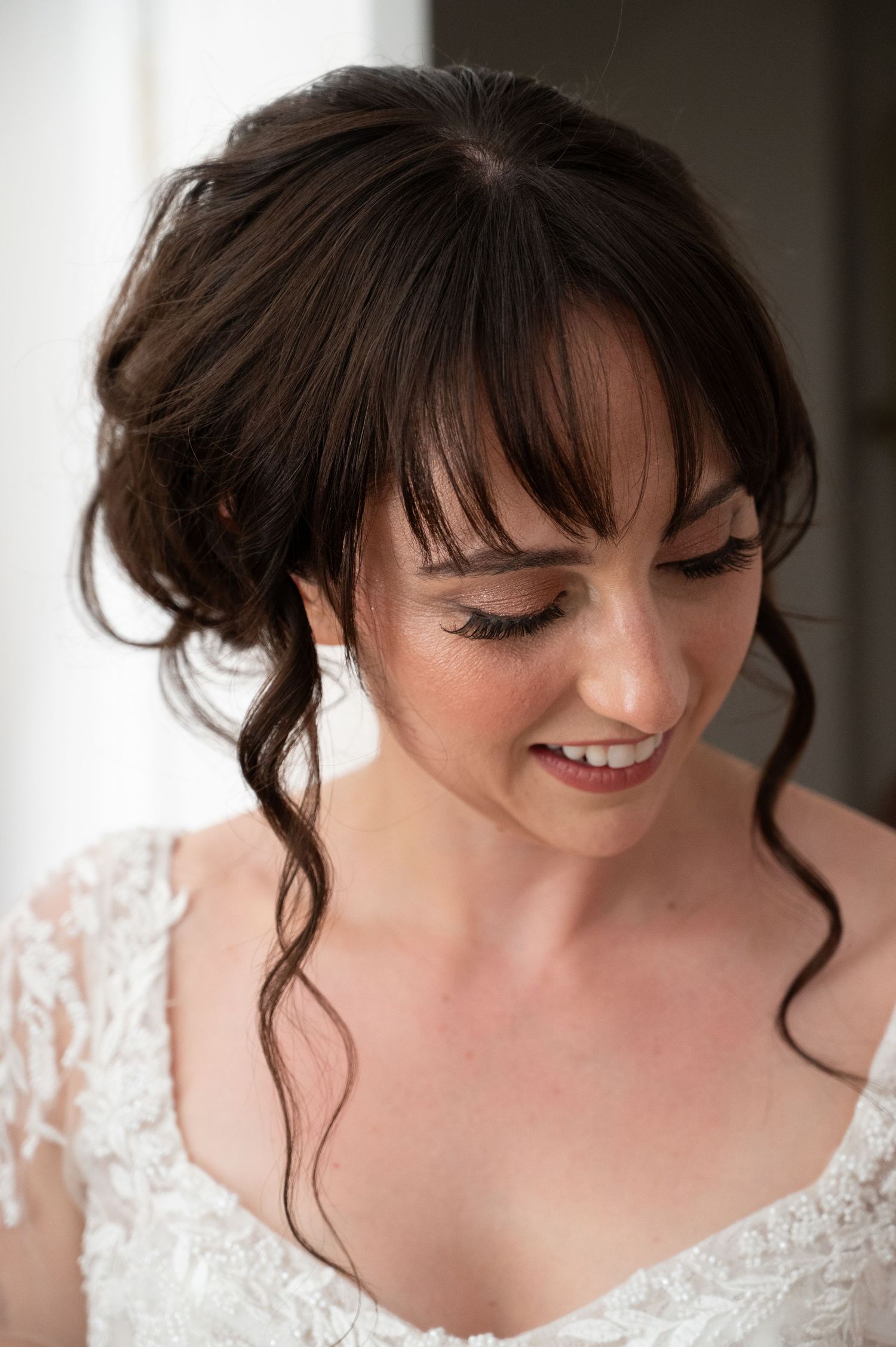 Beautiful bride with dark updo hairstyle and lace wedding dress smiling softly downward.