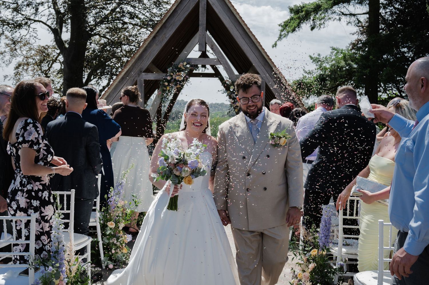 Bride and groom walk down the aisle as guests throw confetti at an outdoor wedding ceremony.