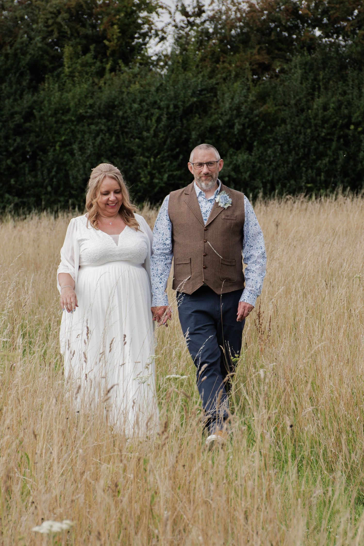 Bride in white dress and groom in brown waistcoat walking hand in hand through tall grass.