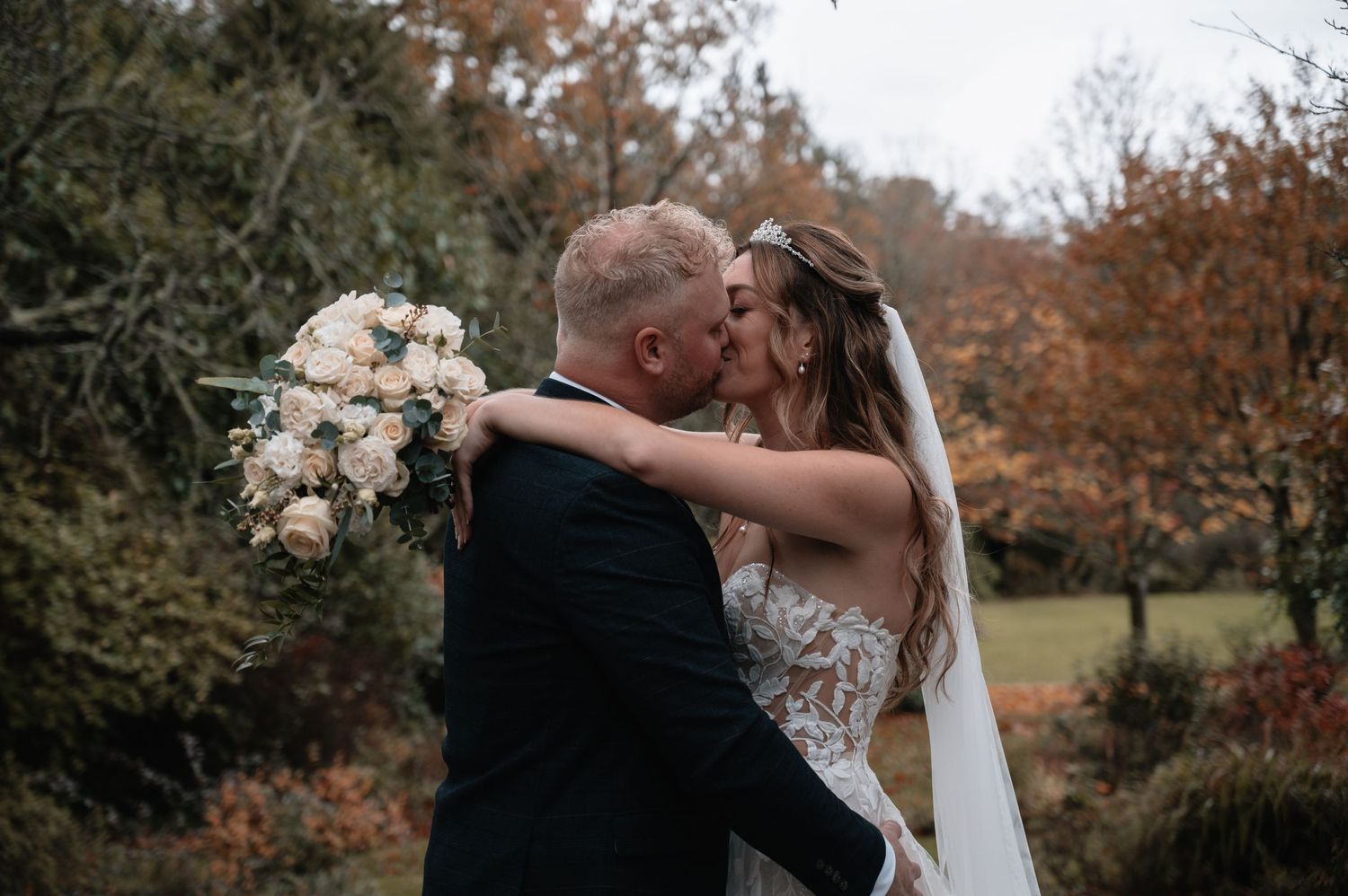 Bride and groom sharing passionate kiss surrounded by vibrant autumn foliage with bridal bouquet.