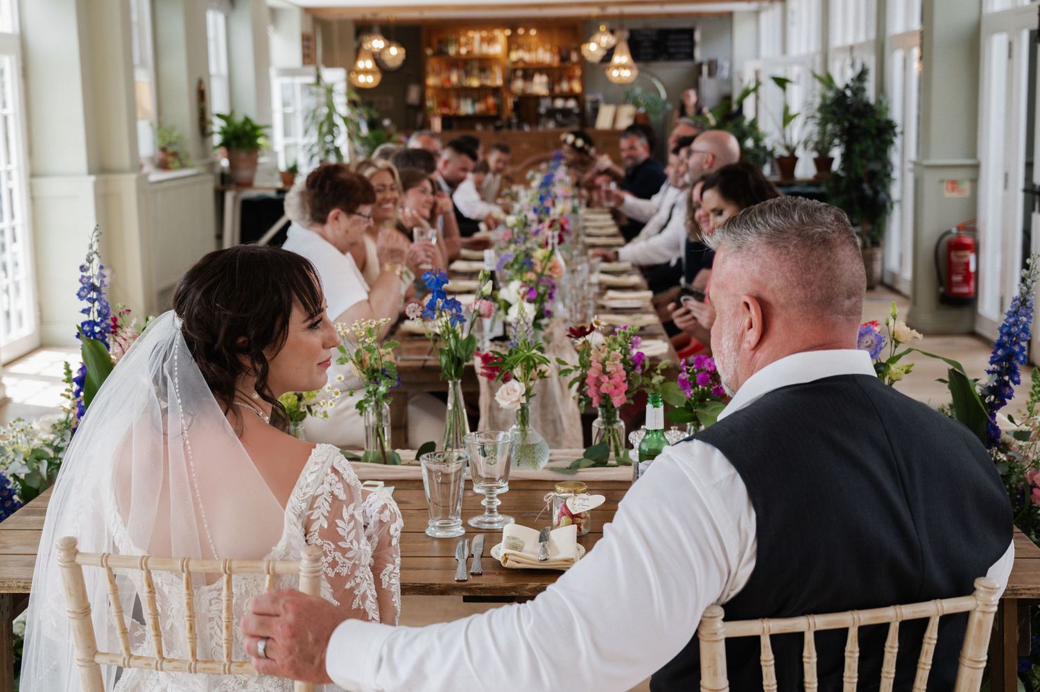 Bride and groom sit at floral-decorated wedding reception table surrounded by celebrating guests.