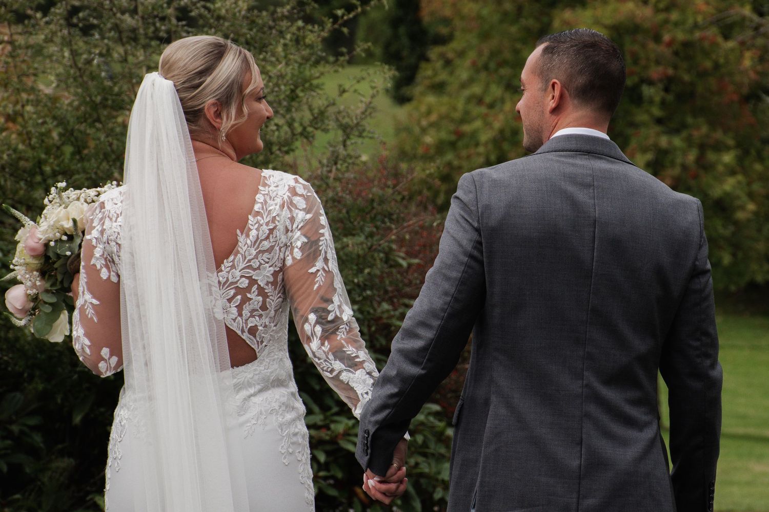 Bride in lace long-sleeve gown and groom in grey suit holding hands walking through garden.