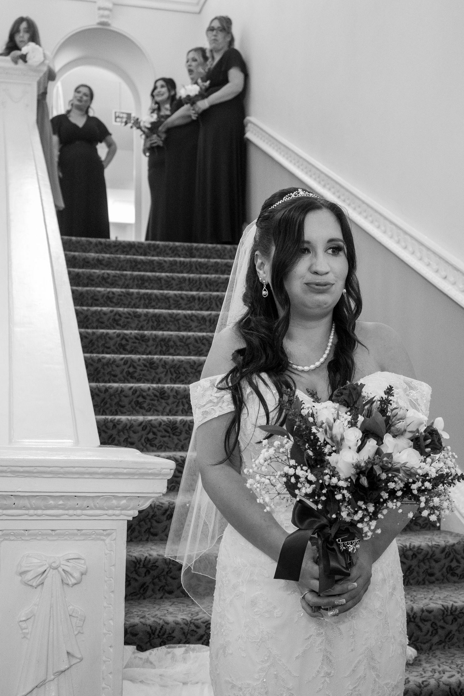 A bride in a lace gown holds a bouquet on a staircase with bridesmaids in black dresses behind her.