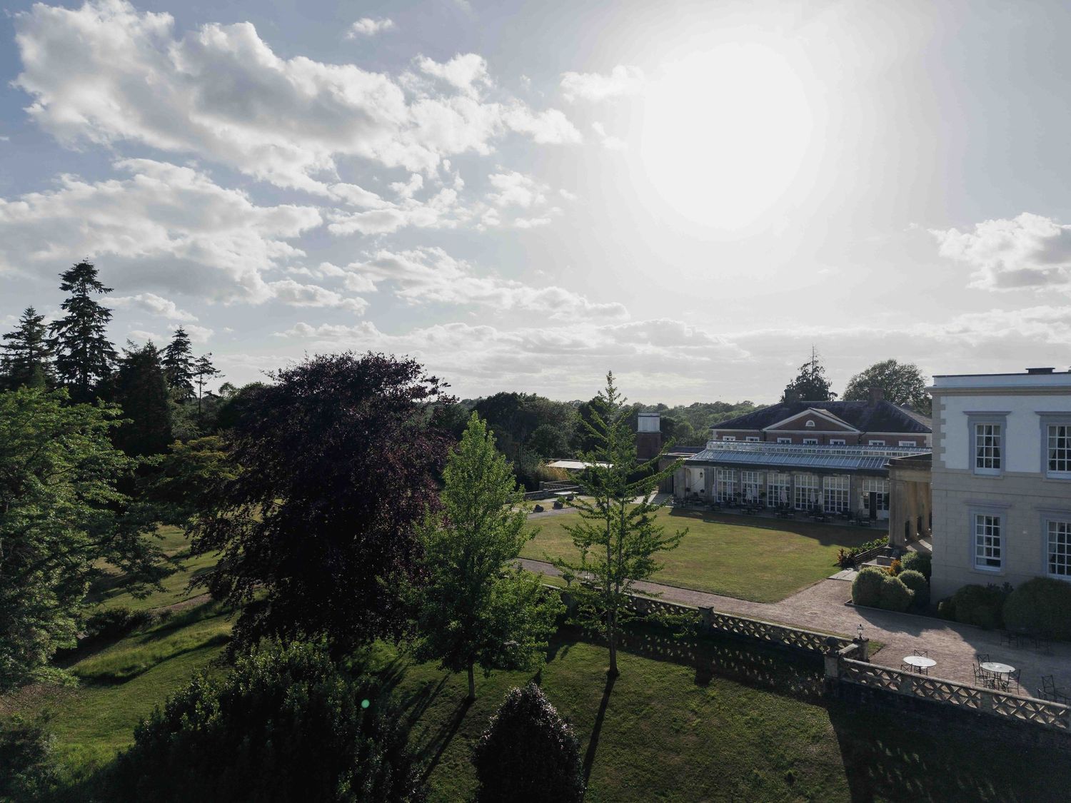 Aerial view of an elegant white country house surrounded by lush trees and manicured gardens on a sunny day. Buxted Park Hotel