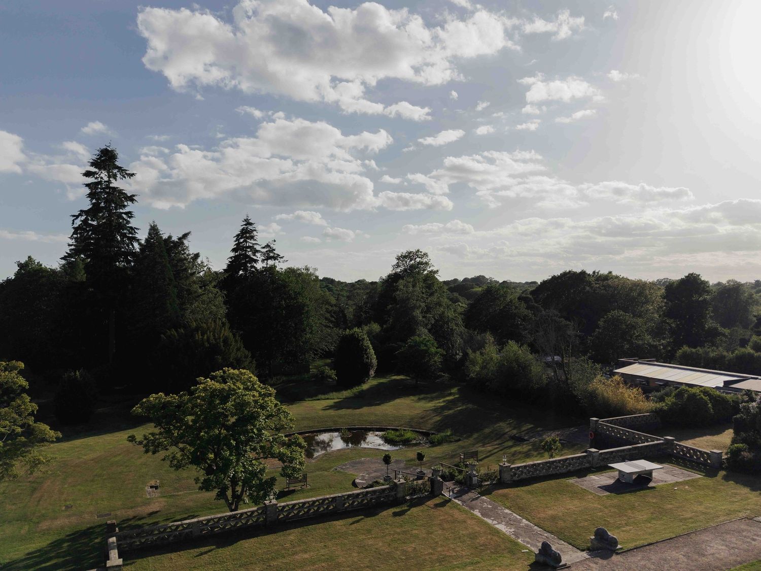 Aerial view of manicured estate grounds with mature trees, a pond, and stone balustrades under a cloudy sky. Buxted Park Hotel