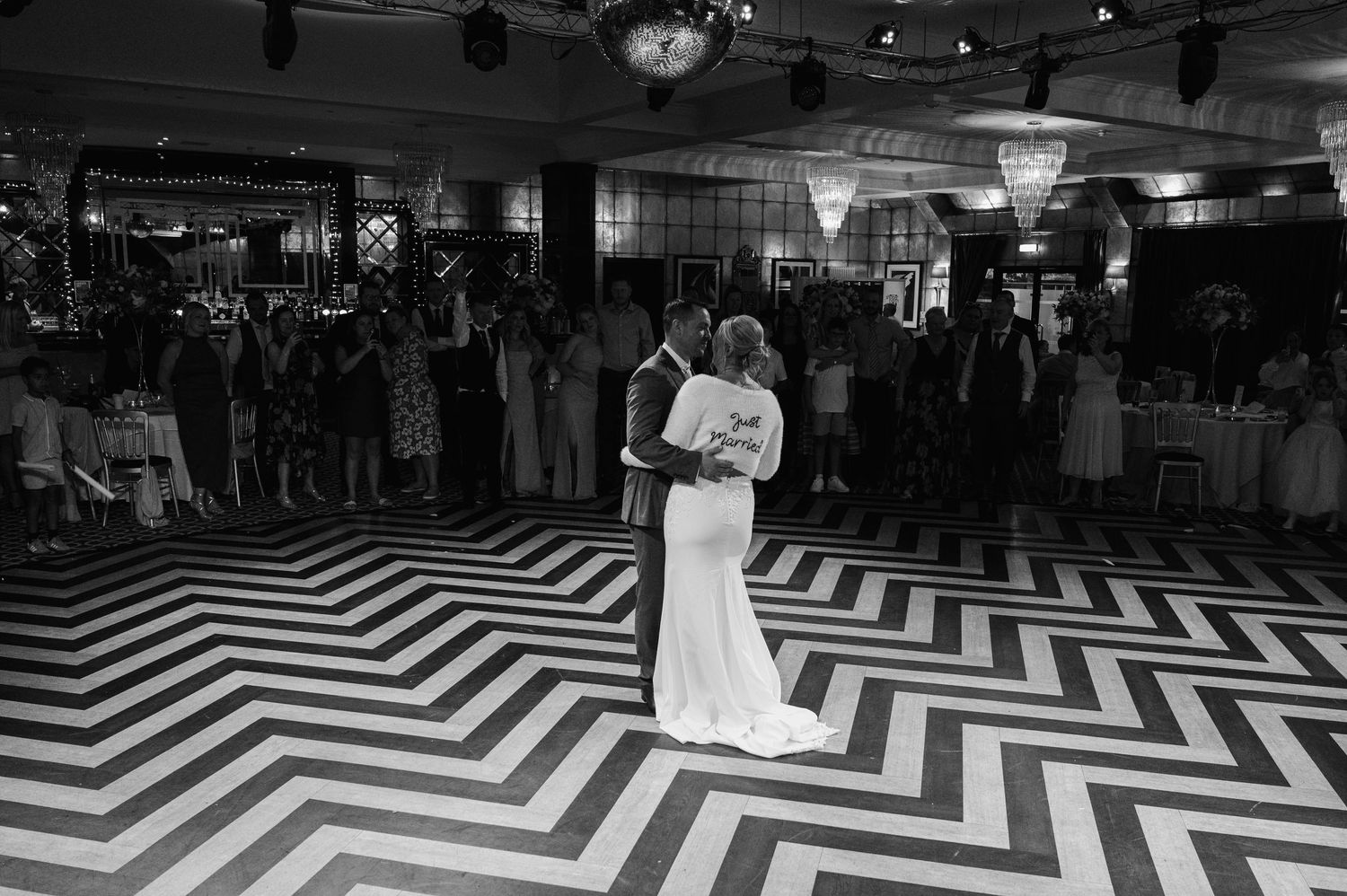 Couple sharing first dance on striking black and white chevron patterned floor surrounded by wedding guests.