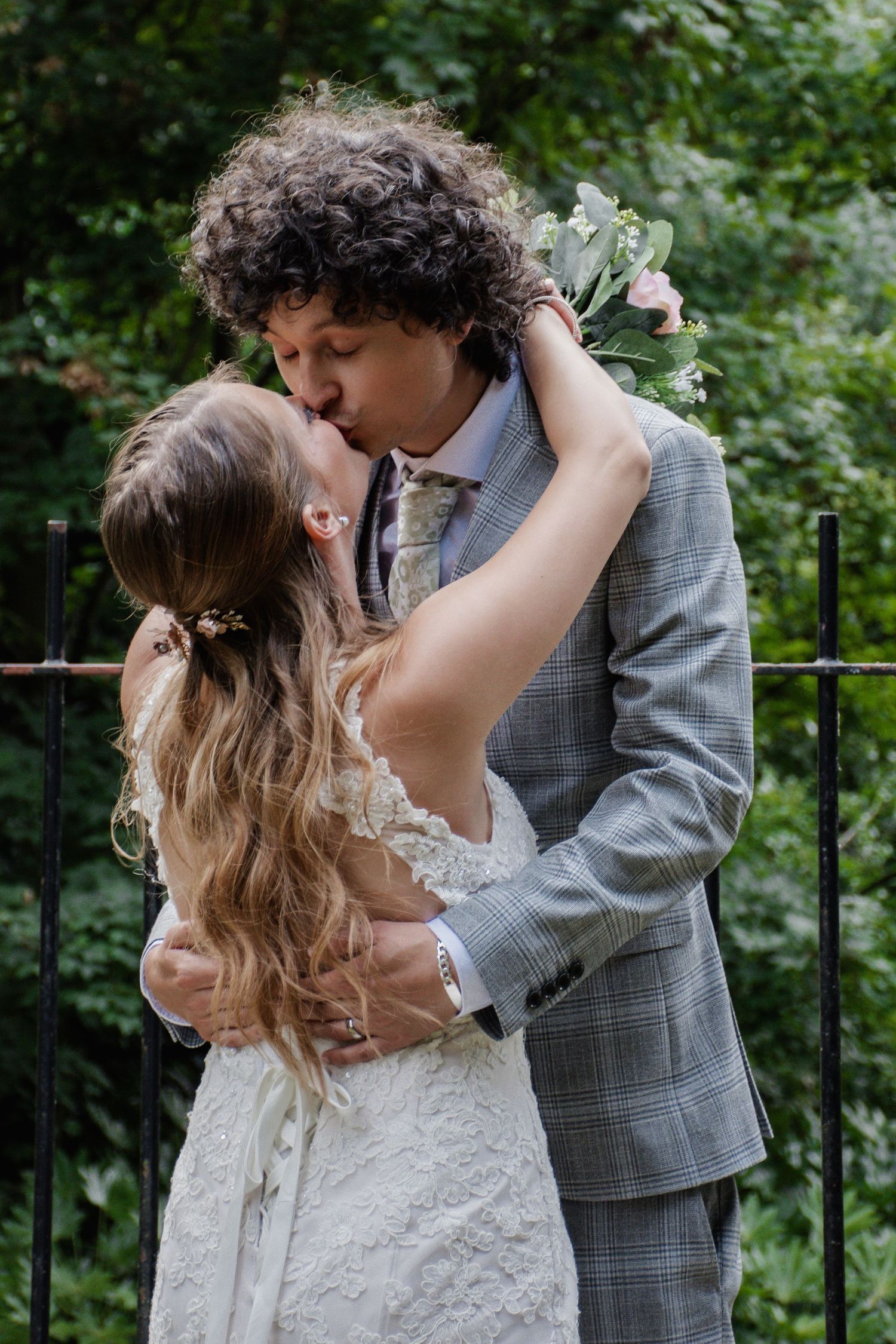 A bride and groom share a romantic kiss while embracing in front of lush green foliage.