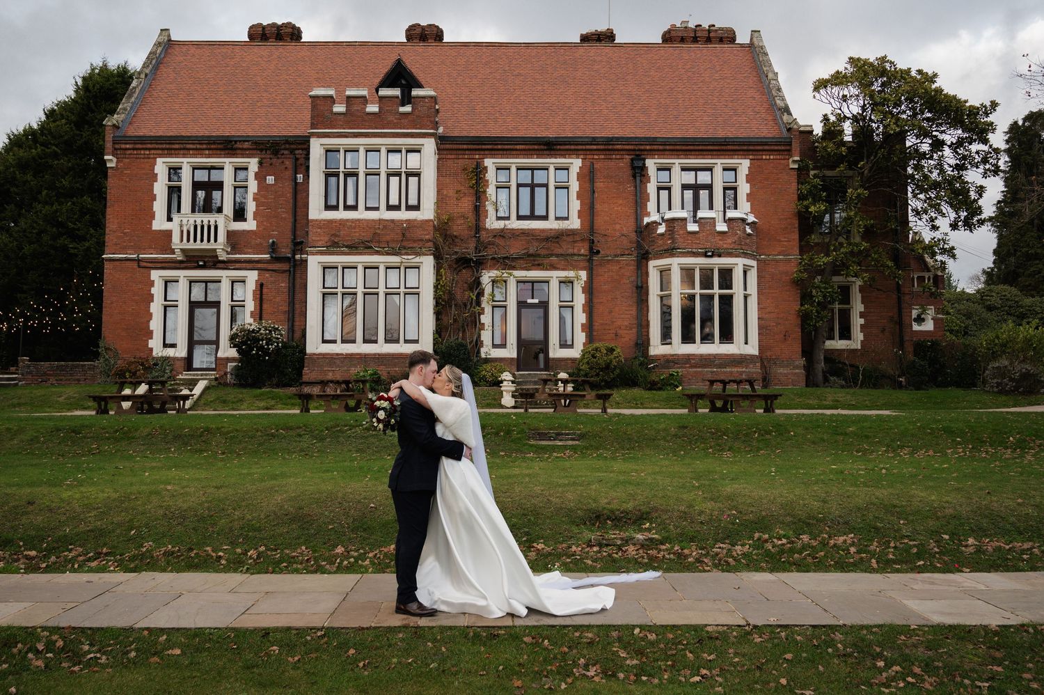 Bride and groom sharing romantic kiss outside elegant red brick English manor house wedding venue.