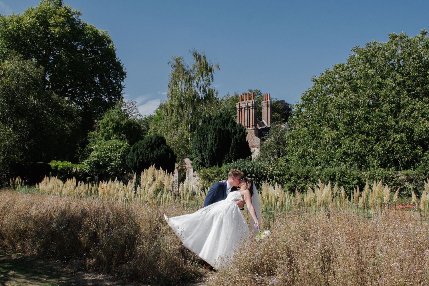 Bride and groom share a romantic kiss in a lush garden setting with historic brick building backdrop on their wedding day.