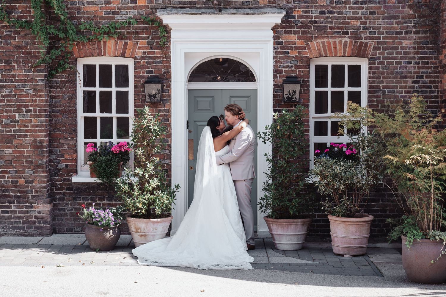 Bride and groom sharing a romantic kiss outside a brick building with potted plants and flowers on their wedding day.