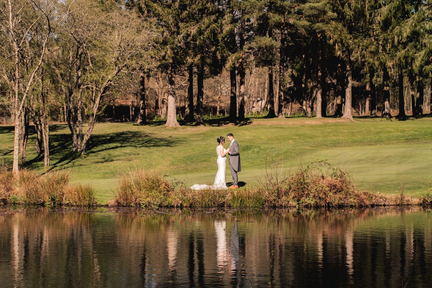 Bride and groom embracing romantically by a reflective lake surrounded by autumn trees and lush greenery.