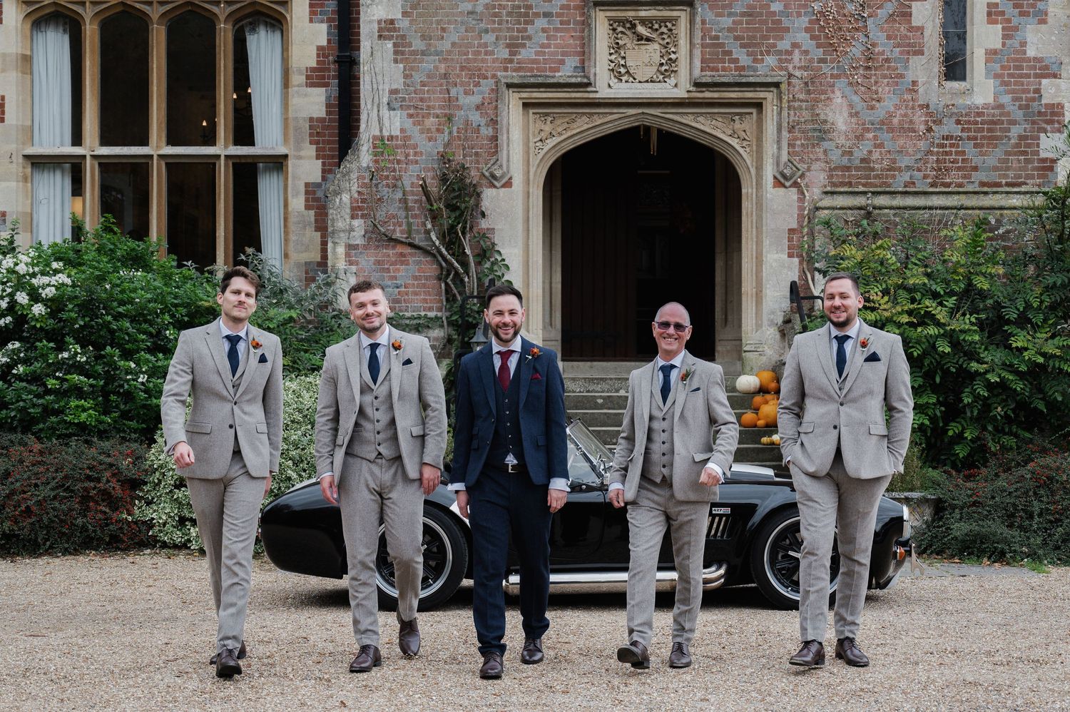 Five groomsmen in grey suits posing confidently in front of a classic car at a grand manor wedding venue.