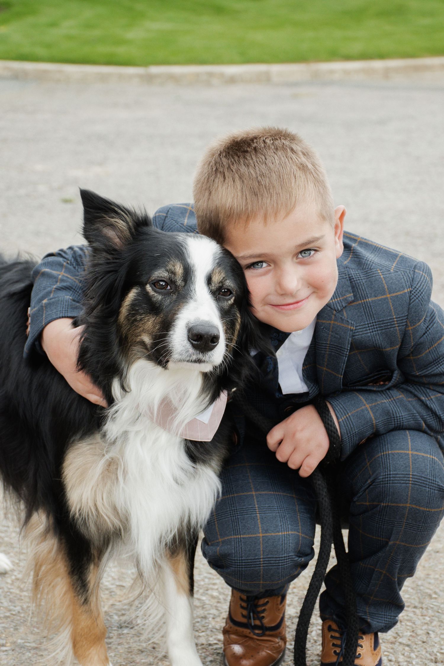 Smiling boy in checked suit hugging black and white border collie at wedding.