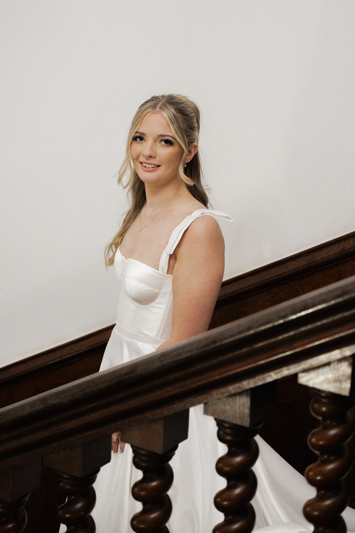 Smiling blonde bride in white satin corset gown posing on ornate wooden staircase in elegant venue.