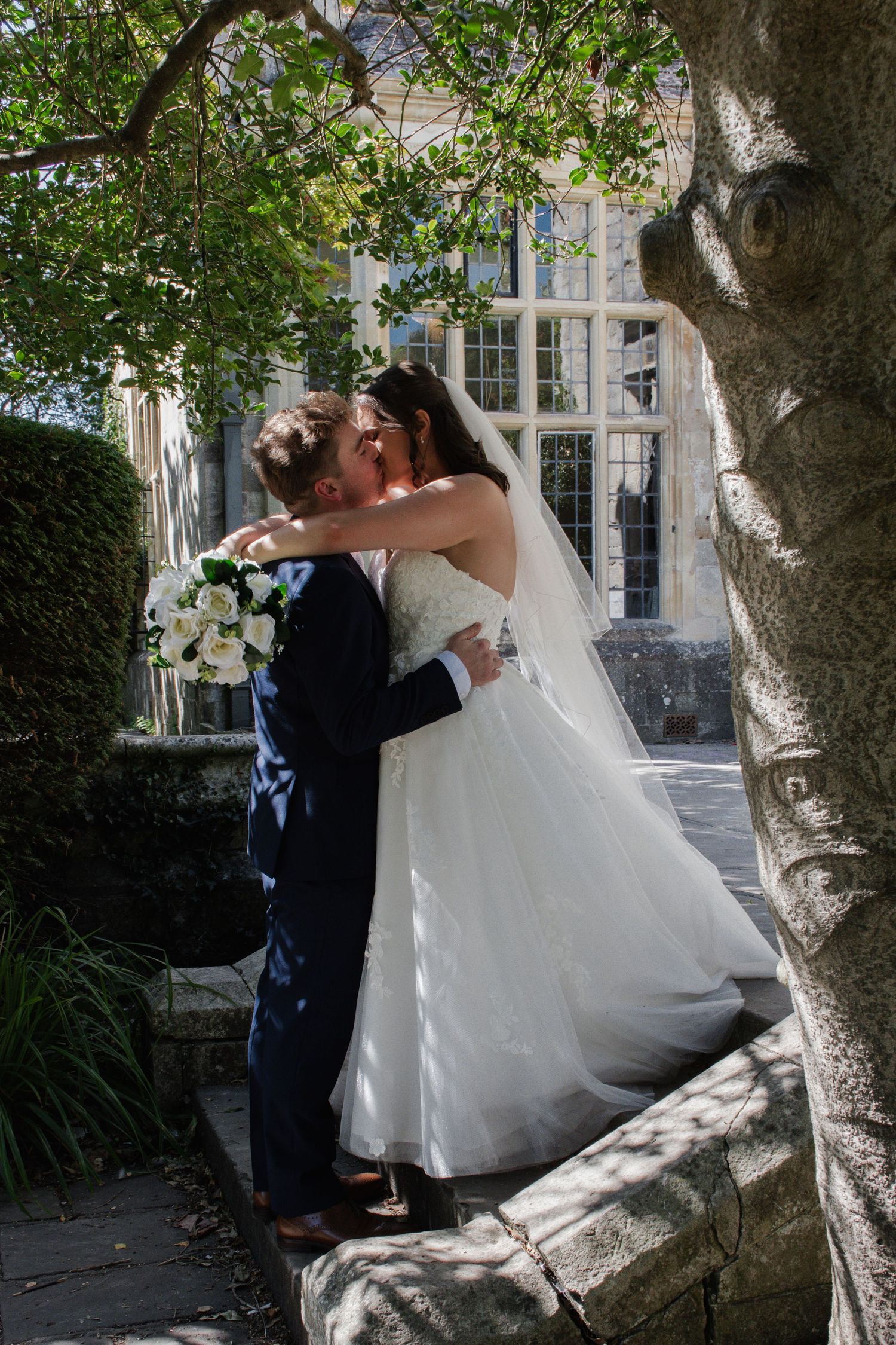 Bride and groom share a tender kiss beneath a sprawling tree outside a historic stone manor house wedding venue.