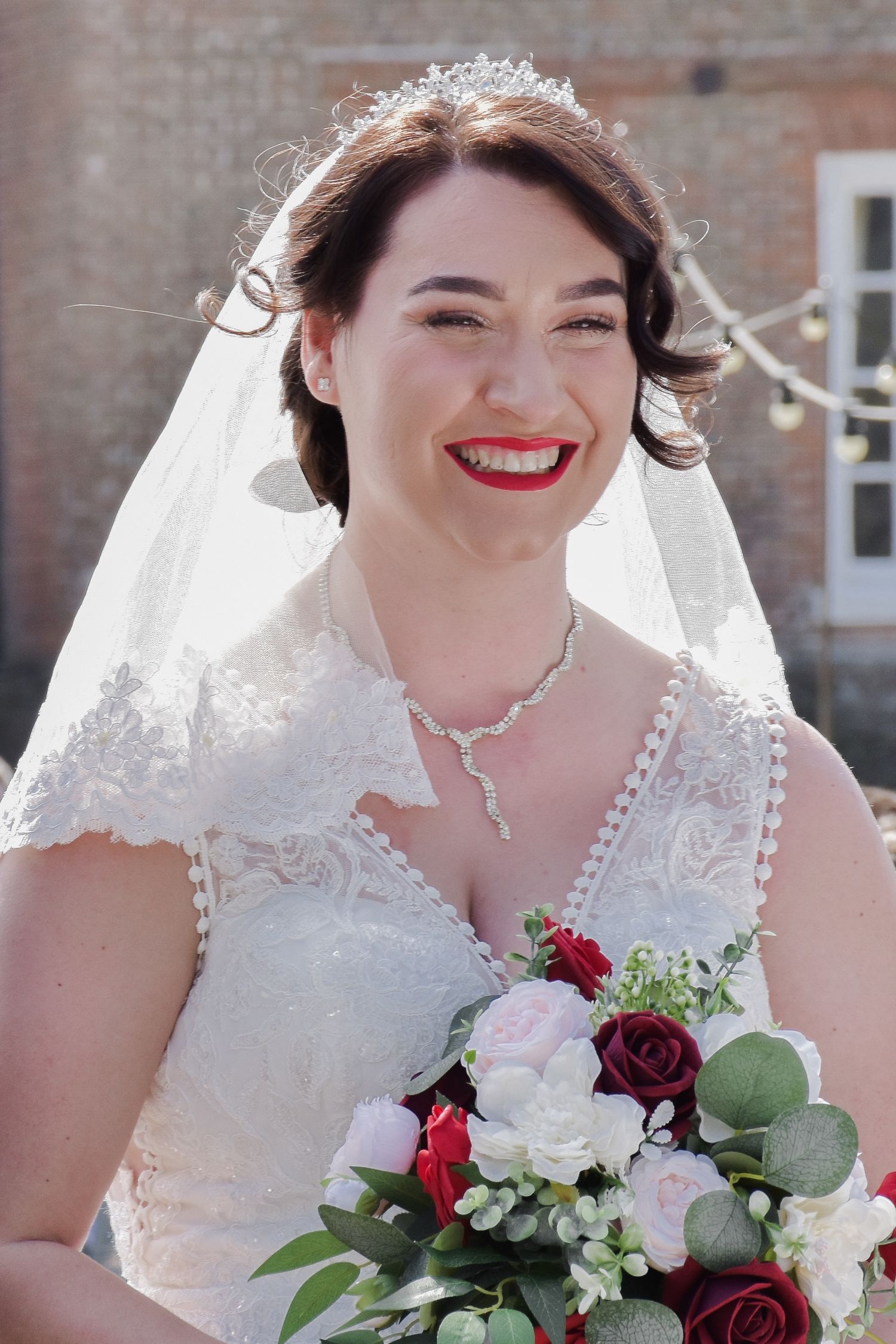 Smiling bride wearing a lace veil and tiara holding a bouquet of red and white roses.