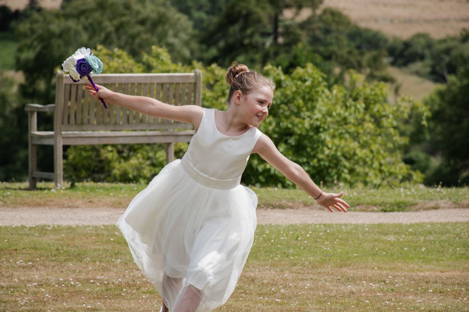 A joyful flower girl in a white dress twirls while holding a bouquet in a sunny garden.