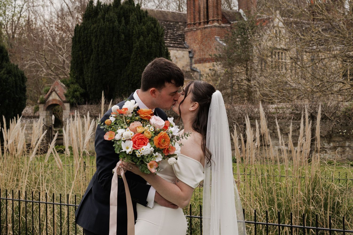 Bride and groom kiss in a garden setting, bride holding a vibrant orange and pink bouquet with ribbon streamers. Lewes registry office.