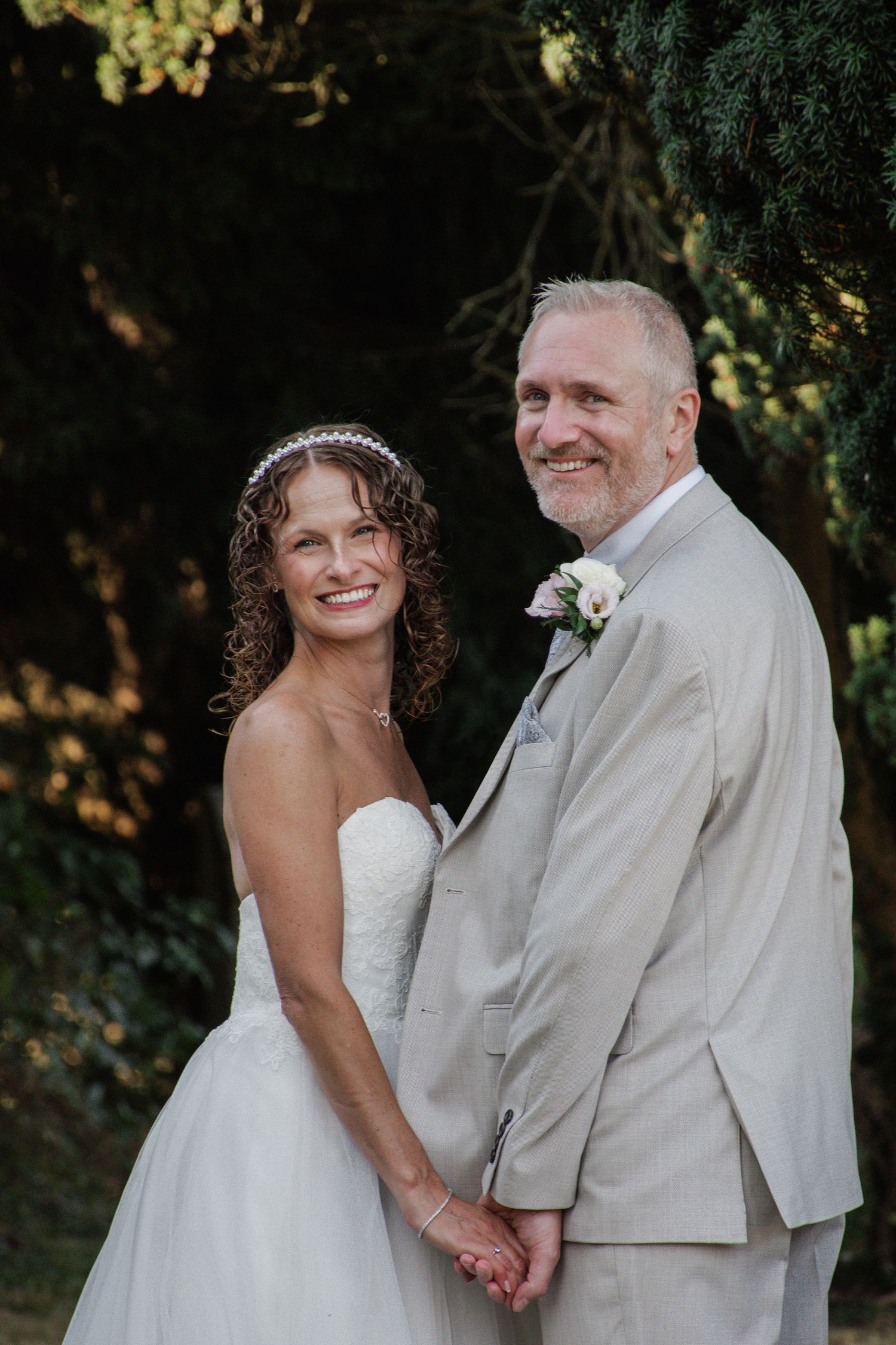 Smiling mature bride and groom hold hands wearing strapless white gown and beige suit in a lush garden setting.