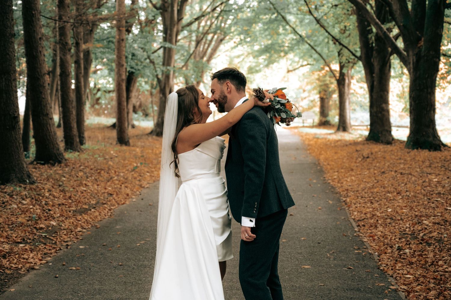 Bride and groom sharing a romantic kiss on an autumn tree-lined path covered in fallen orange leaves.