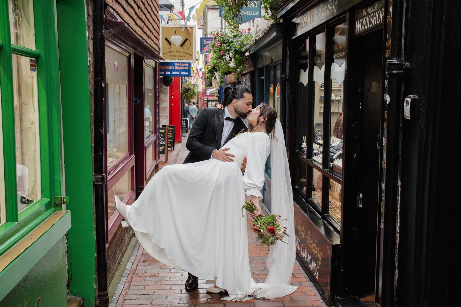 Groom dips bride for a kiss in a quaint cobblestone alley, her white gown flowing as she holds a red bouquet.