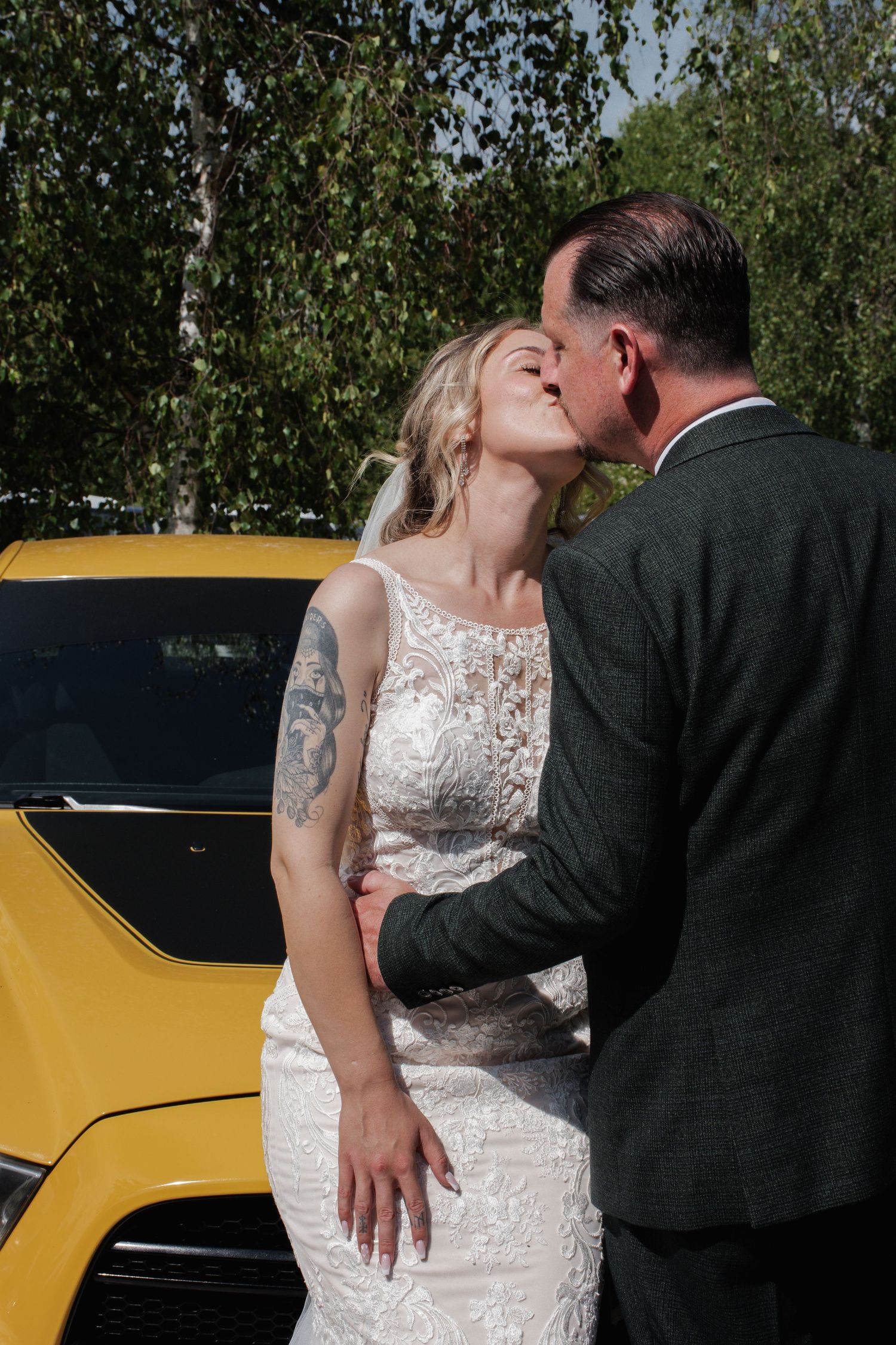 A tattooed bride and groom share a kiss in front of a bright yellow car surrounded by birch trees.