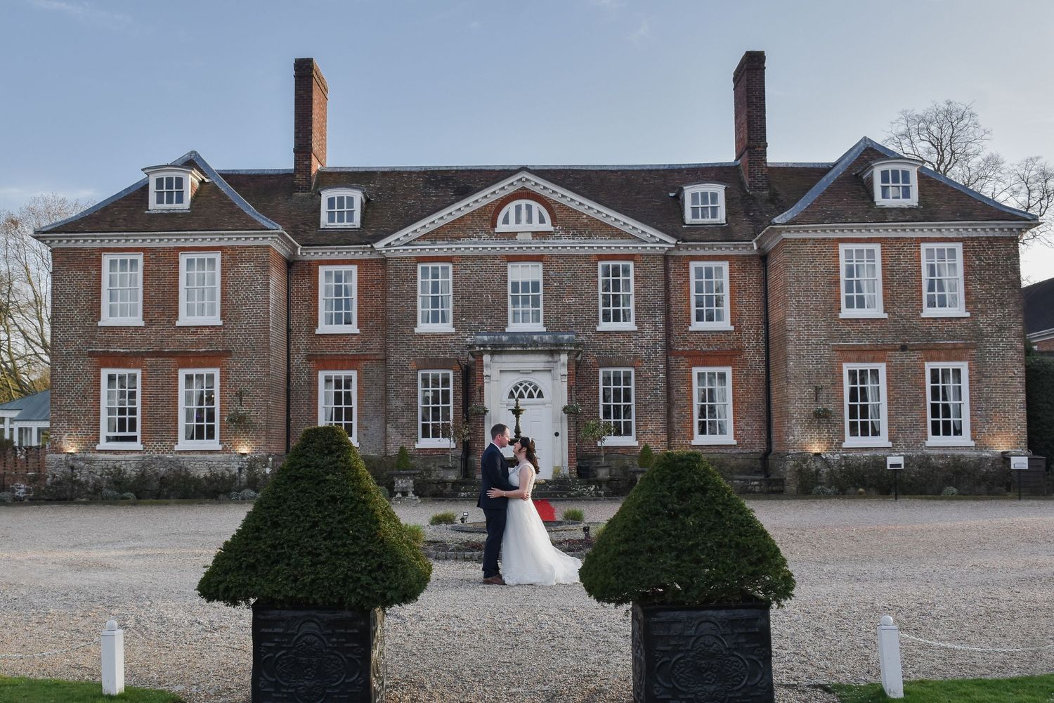 A bride and groom pose together in front of an elegant Georgian brick country house wedding venue. Chilston Park Hotel