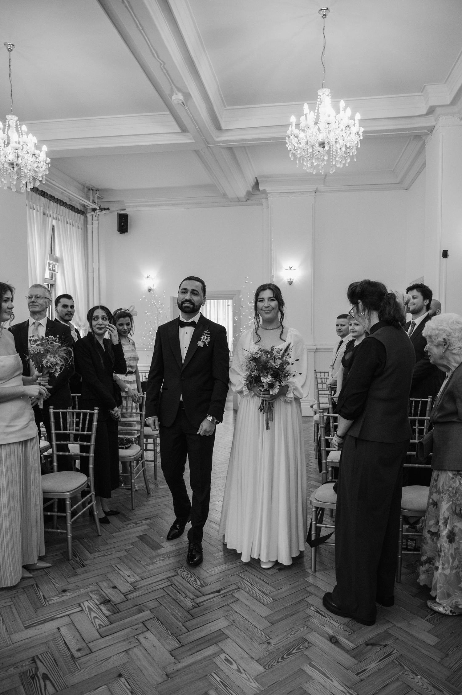 Black and white photo of bride and groom walking down the aisle in an elegant chandelier-lit wedding ceremony hall.