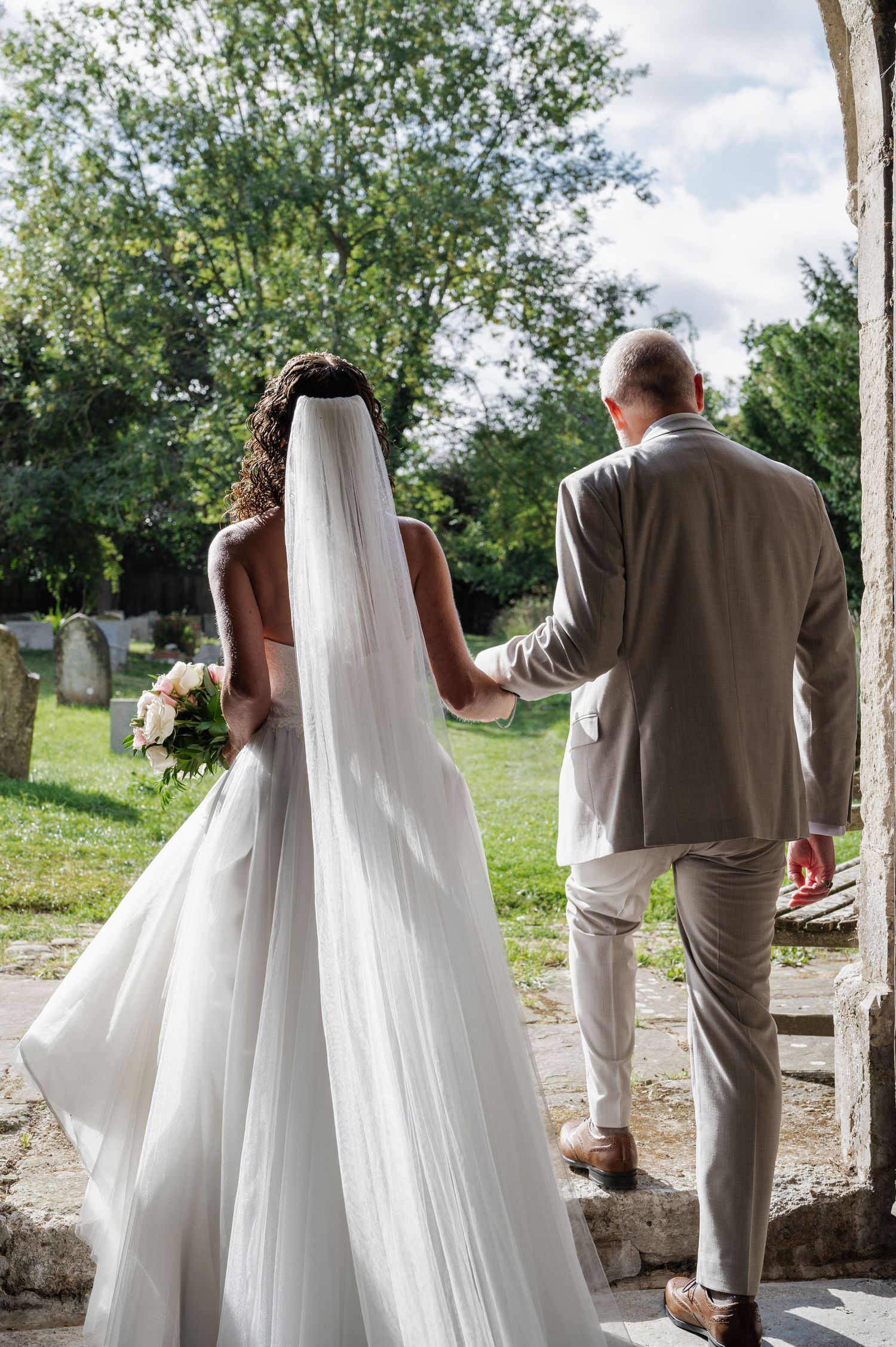Bride in white gown and veil walks hand-in-hand with groom in grey suit toward church entrance.