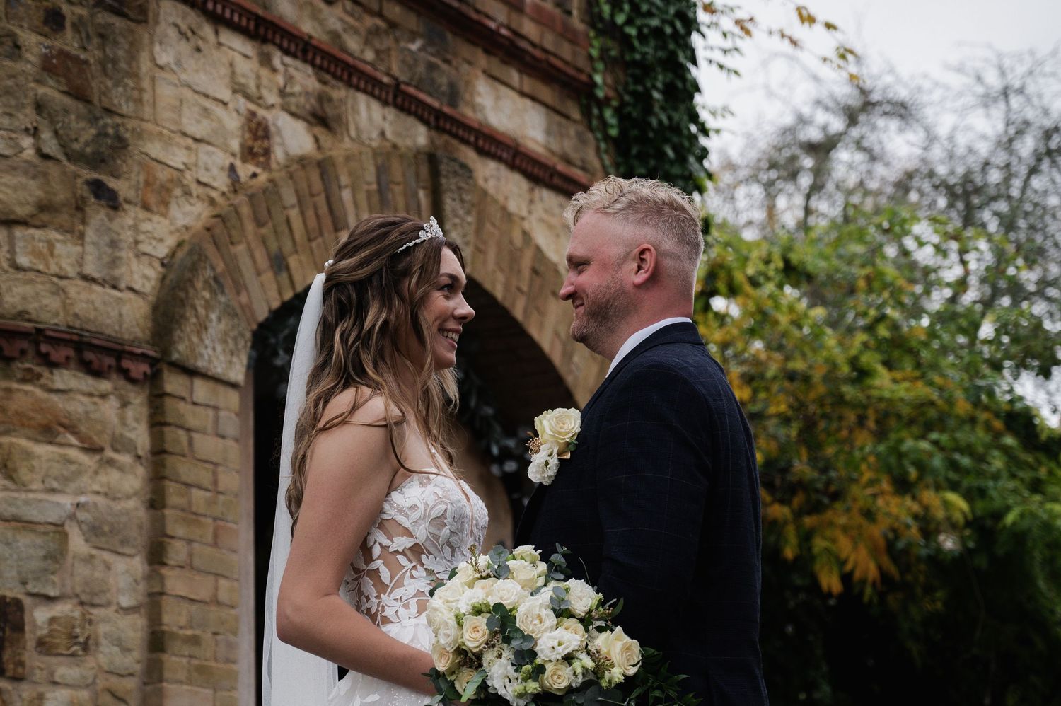 Bride in floral gown and groom in navy suit smile at each other beside a stone church arch.