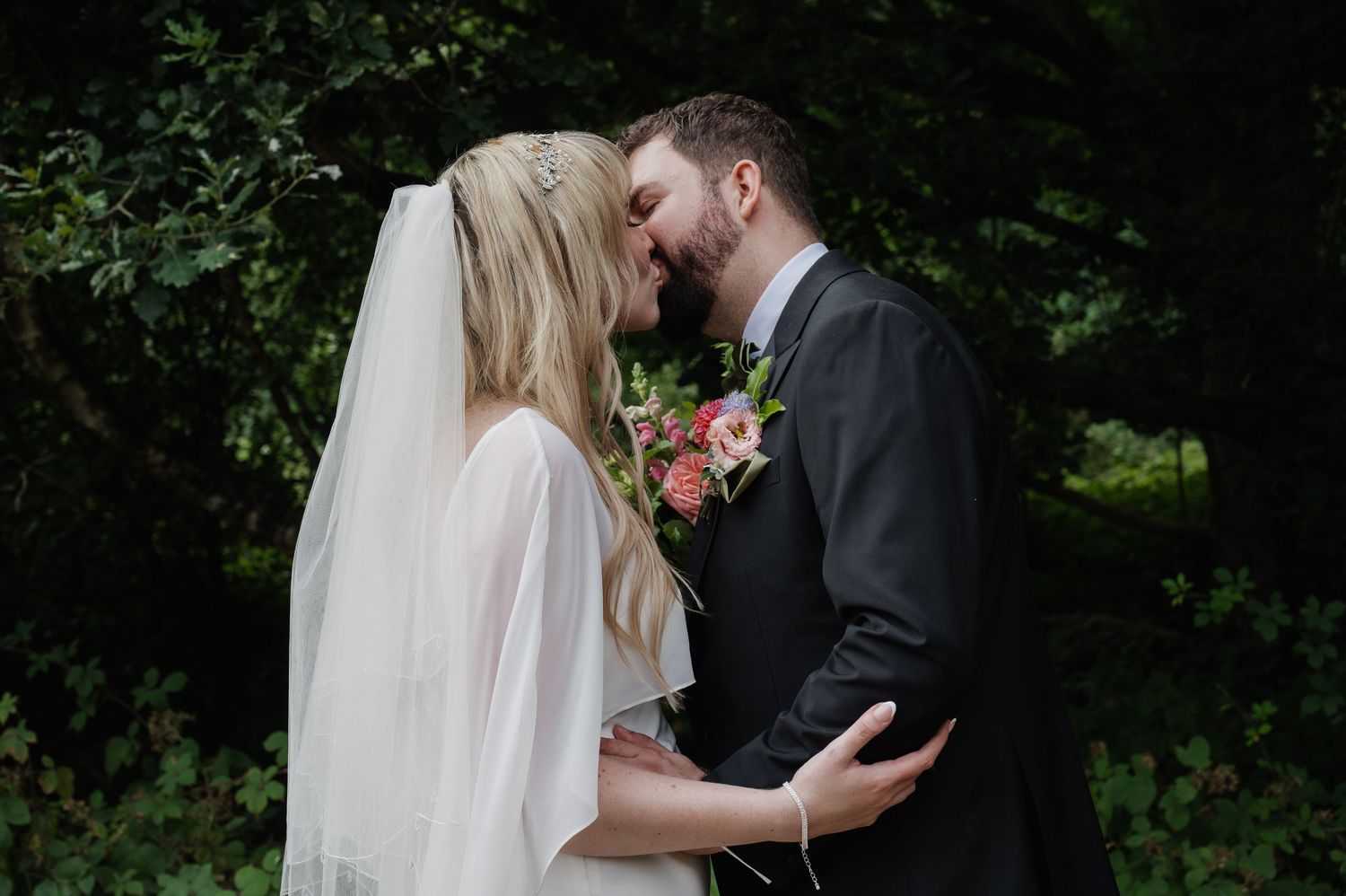 Bride and groom sharing a romantic first kiss, bride wearing a veil and holding a pink floral bouquet outdoors.