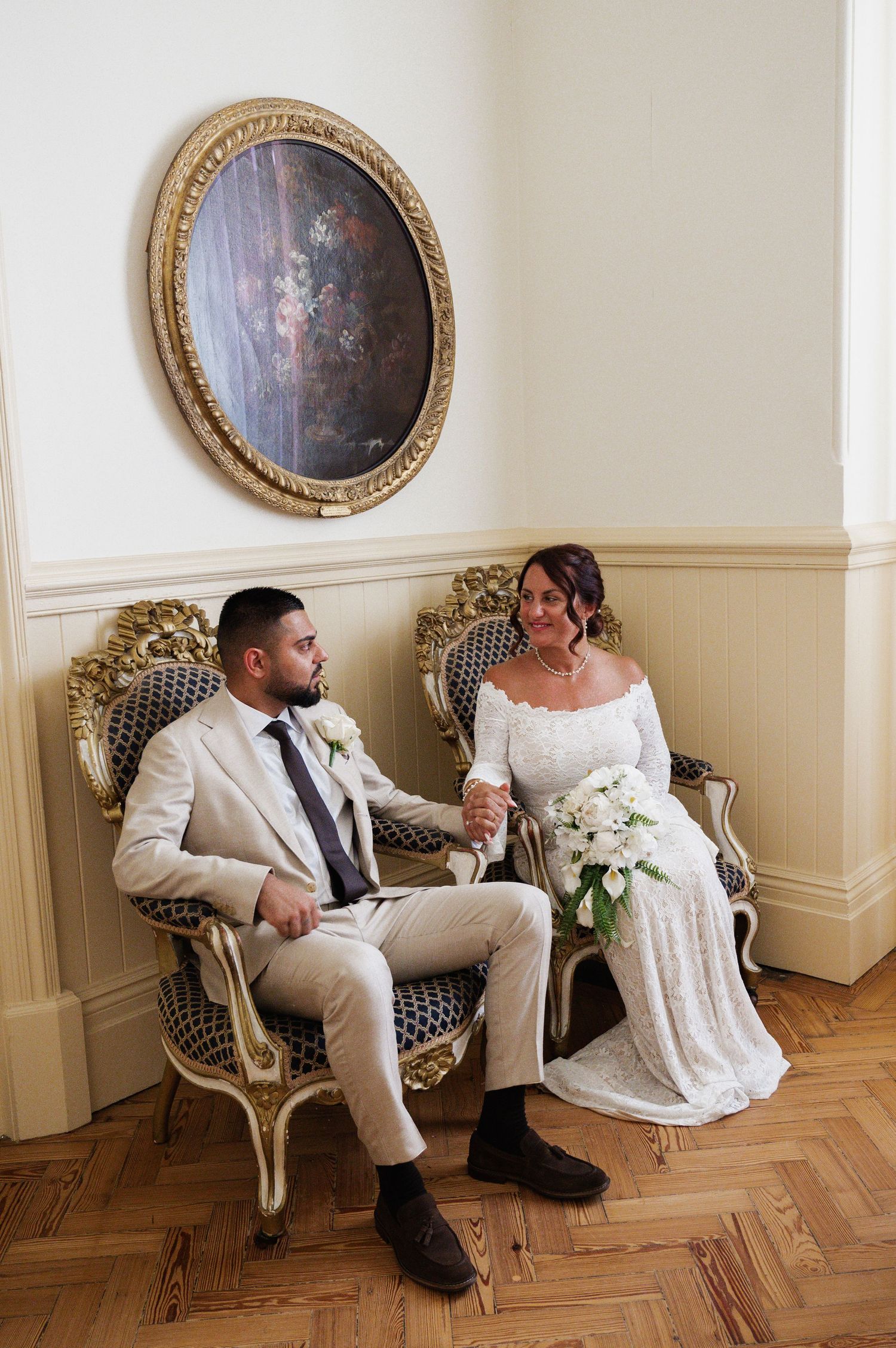 Wedding couple seated in ornate gold chairs holding hands in elegant room with classical painting.